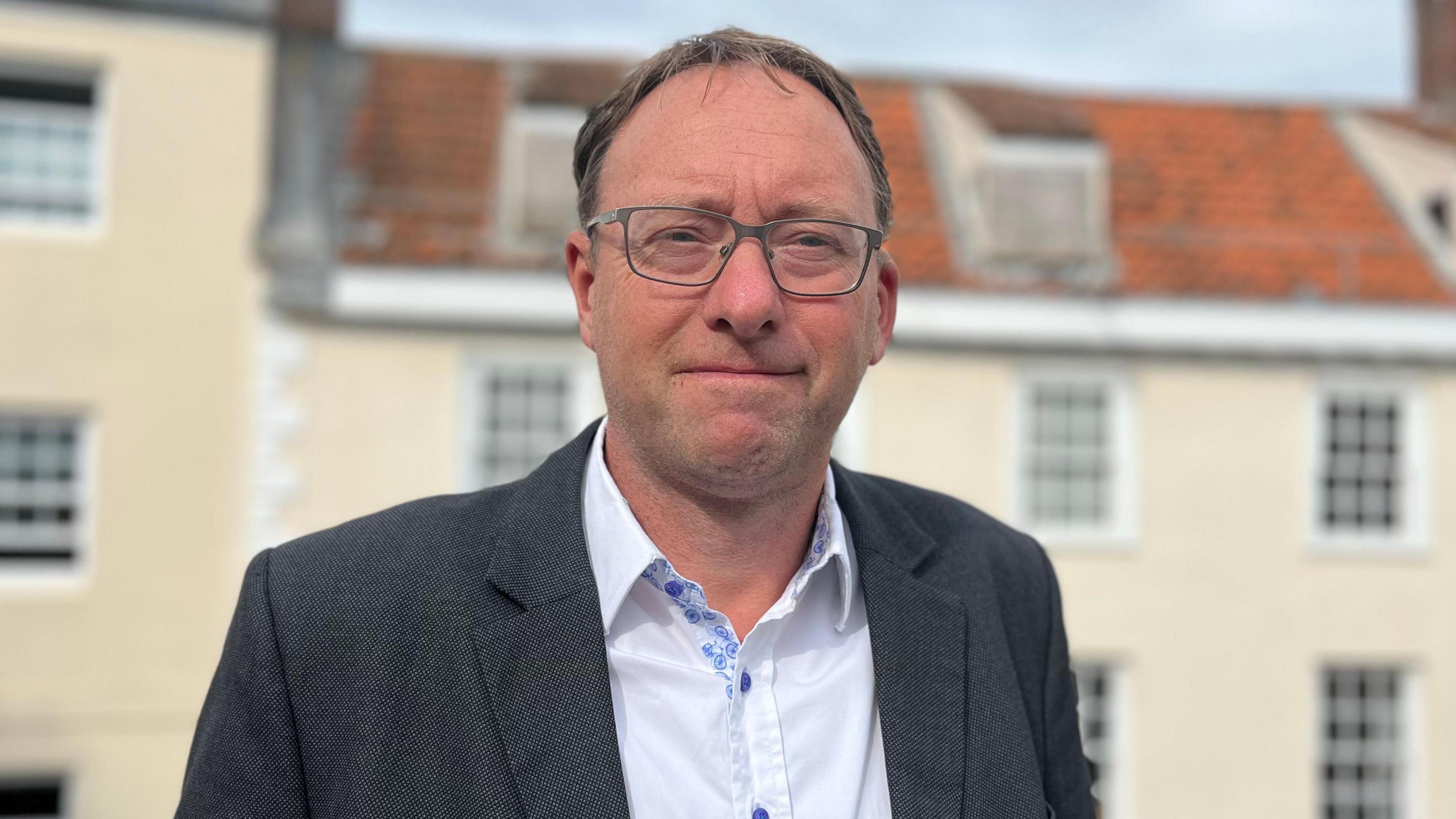 Pierre Blampied, a man in a suit with glasses and a shirt with the top button undone smiles towards the camera.