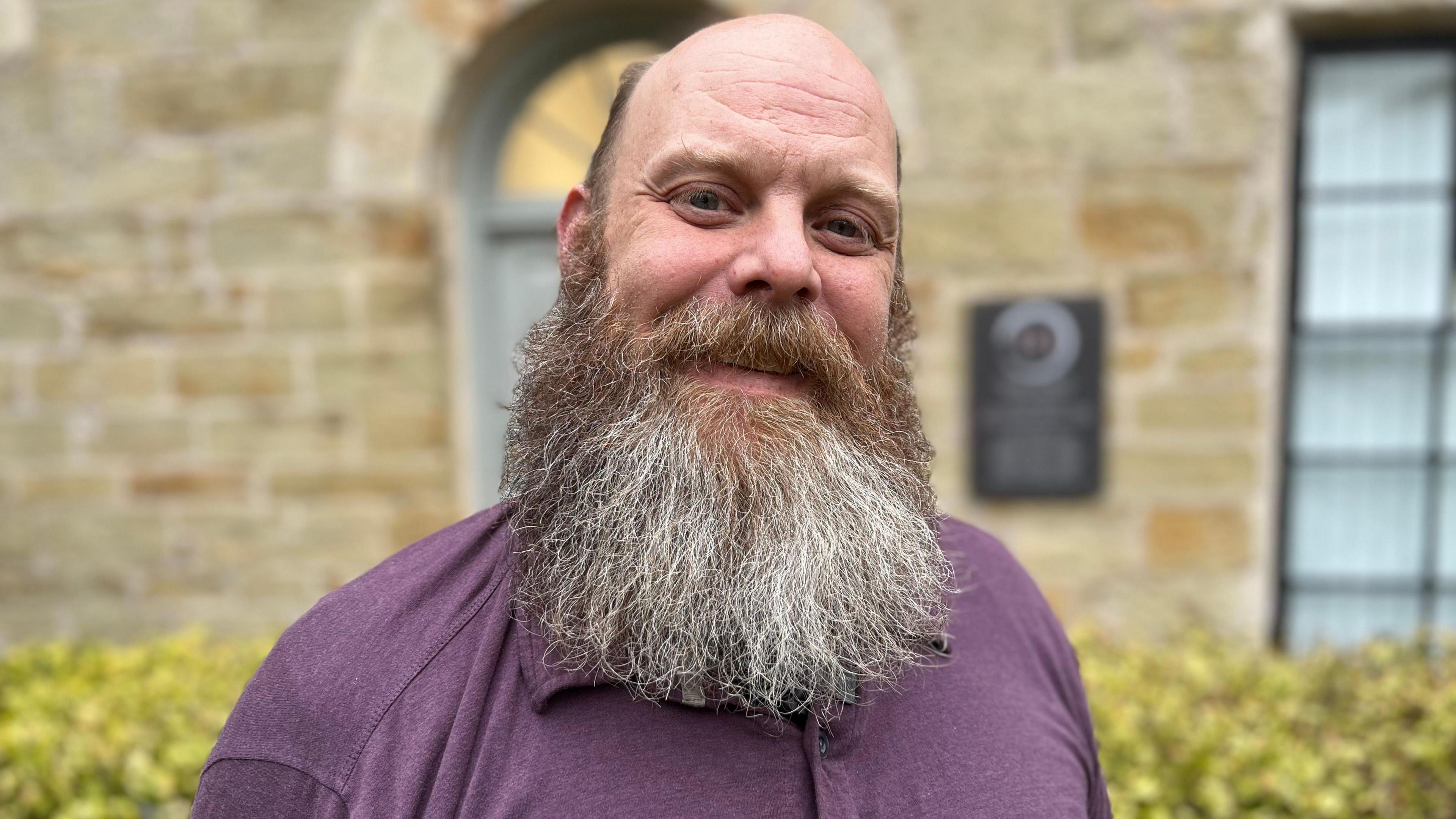 Bobby Inman has a beard and is wearing a purple top. He is stood outside and there's a building and hedge behind him.