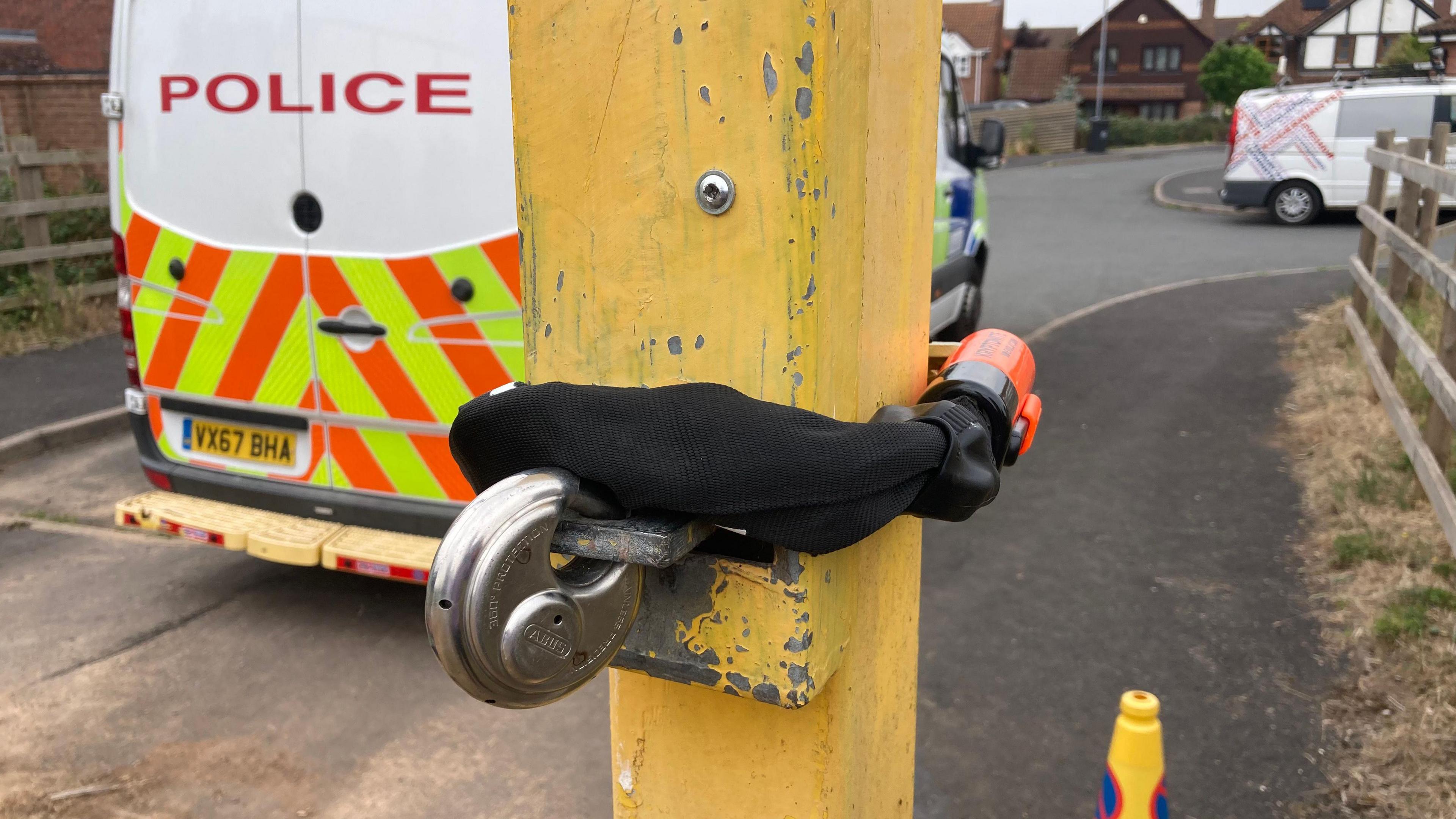 A black bicycle lock is wrapped around a yellow metal gate post. A police van is parked in front of the entrance. 