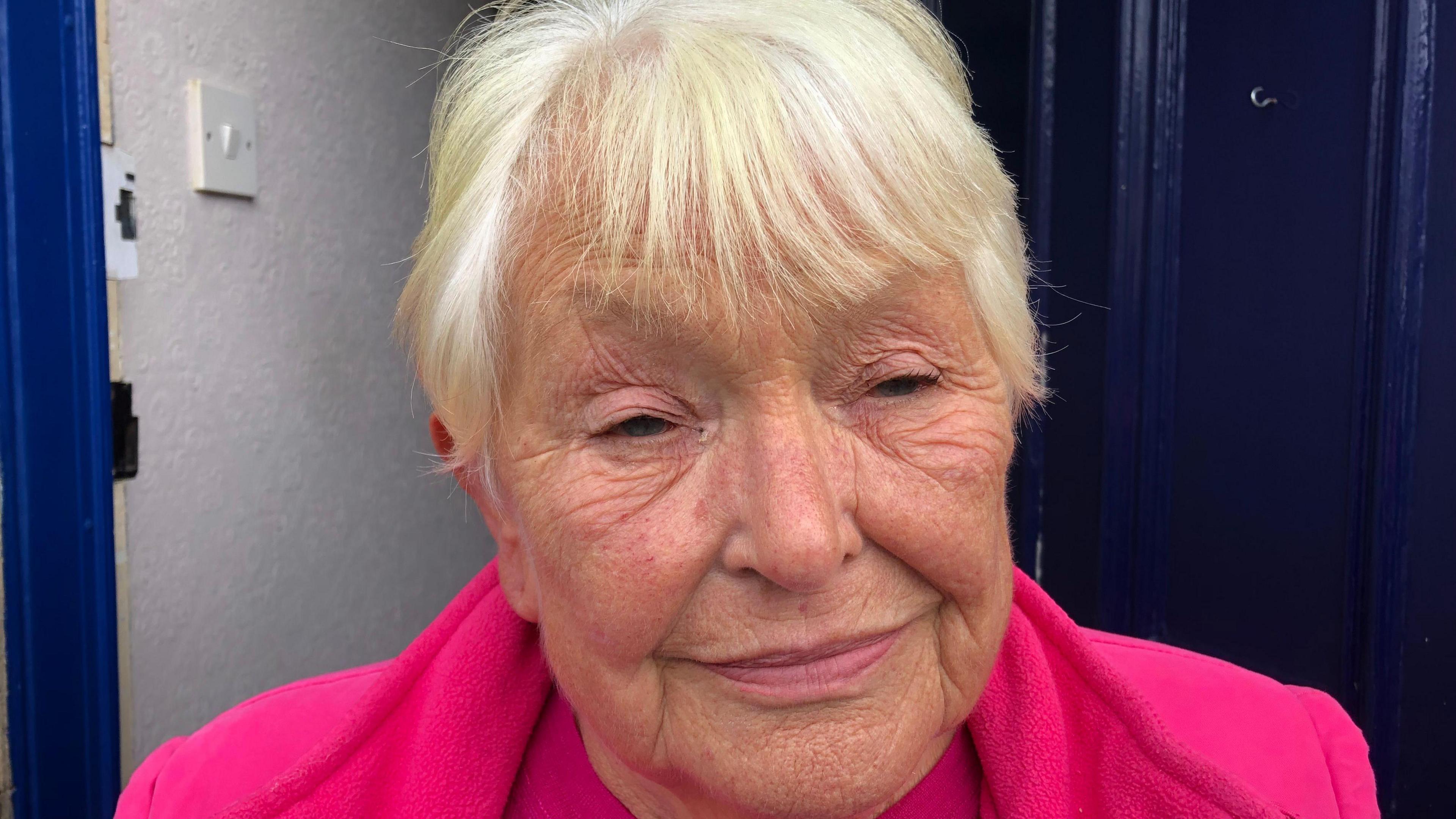 Jean Smith, 74 with white short hair and a pink top with matching fleece in the doorway of her home in Ramsey.