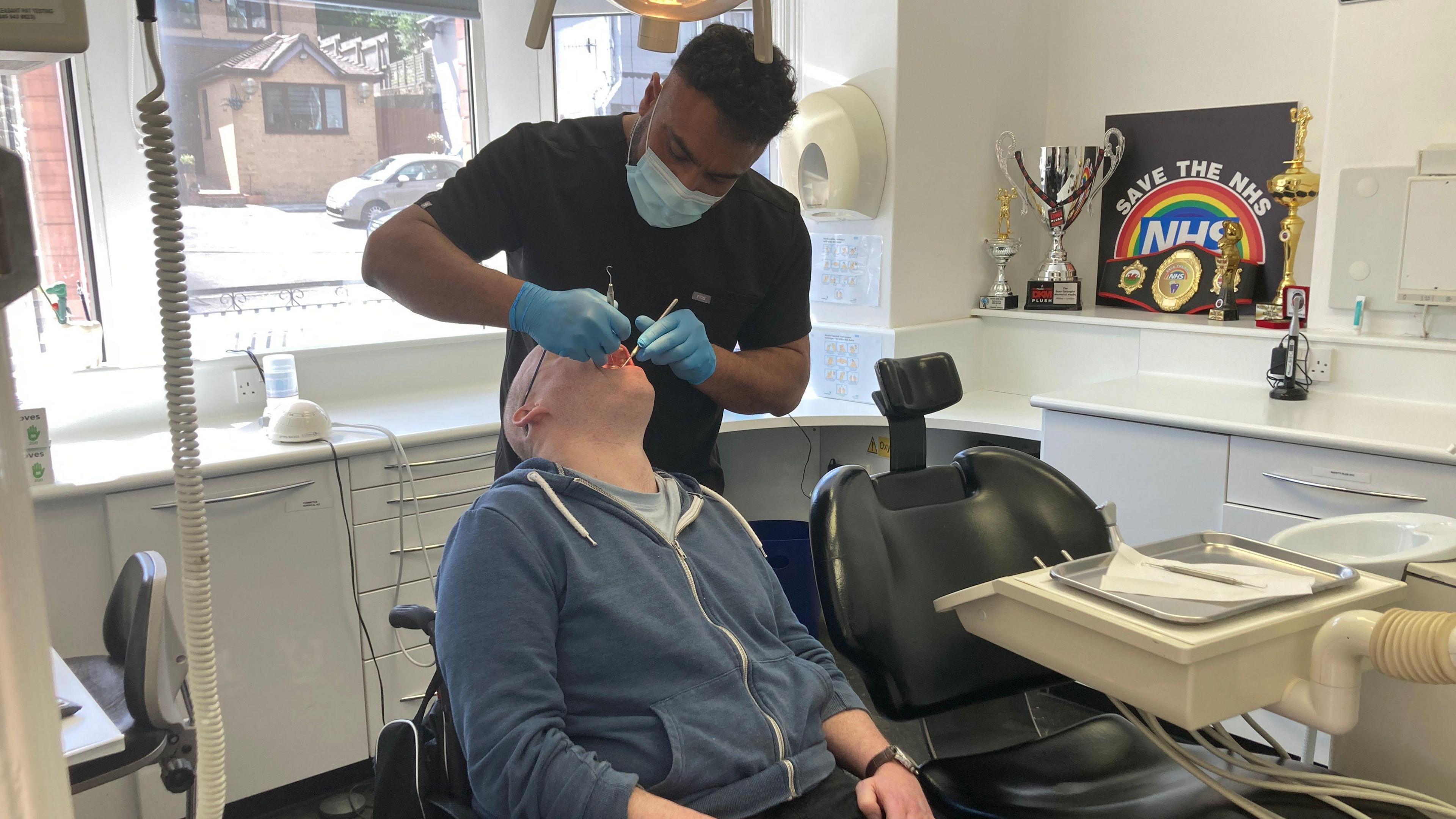 A man in a blue hoodie has his mouth open and is being examined by a dentist, wearing a mask and gloves and a black top. On the wall of the examination room is a sign with 'save the NHS' and a number of trophies and boxing belt.