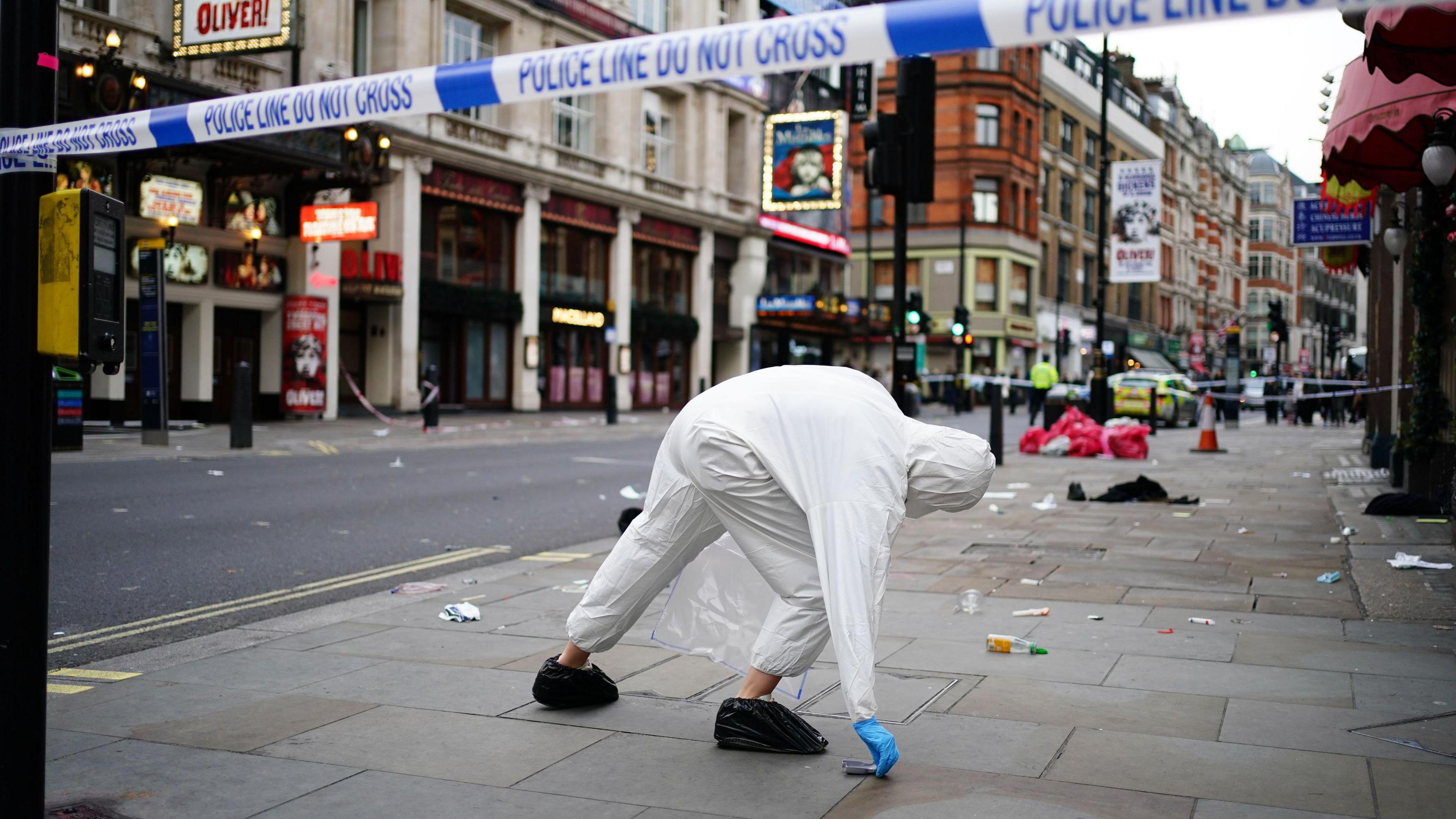 A street closed by police cordons on both ends. A forensic officer in while overalls is picking up an item from the pavement. 