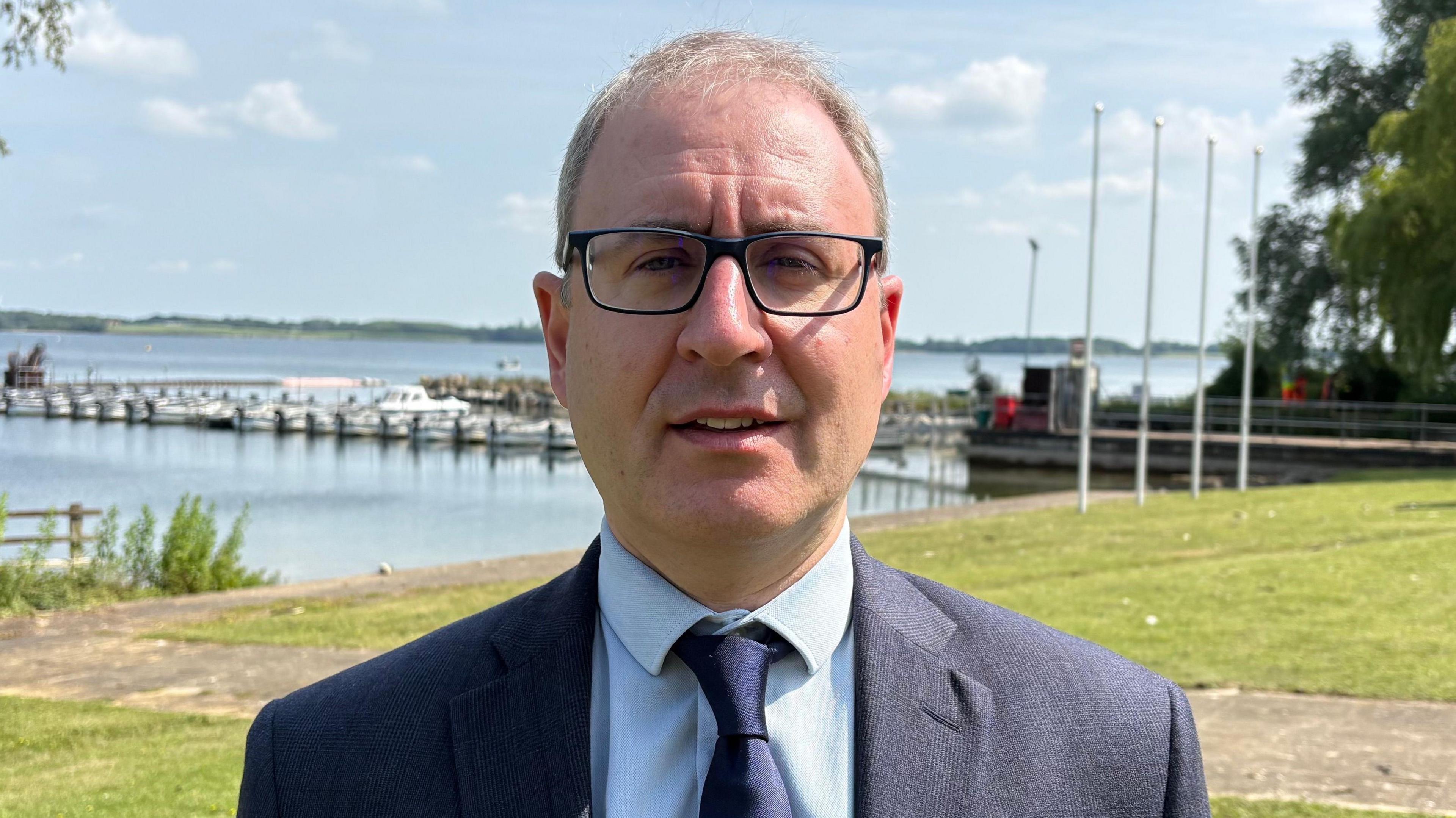 Geoff Darch looking direct to camera. He is standing outside on a sunny day. In the background is a grassy area and some concrete pathways that lead down to the banks of a large lake; some small boats can be seen in the background to the left moored to a pontoon. Mr Darch has greying, short cut hair and is wearing black rimmed, rectangular glasses. He is wearing a dark blue blazer and tie and a light blue shirt.