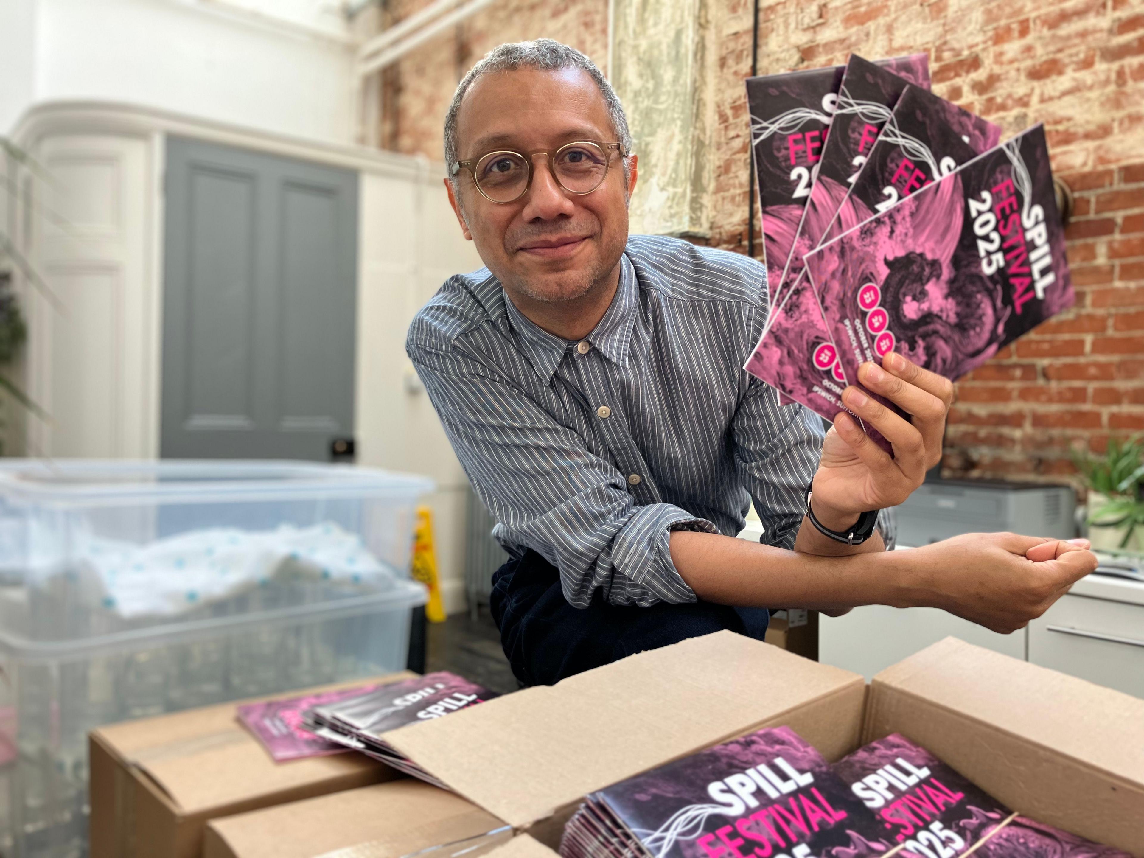 Robin Deacon hold several purple brochures in his hand as he leans on a stack of boxes also containing the programmes