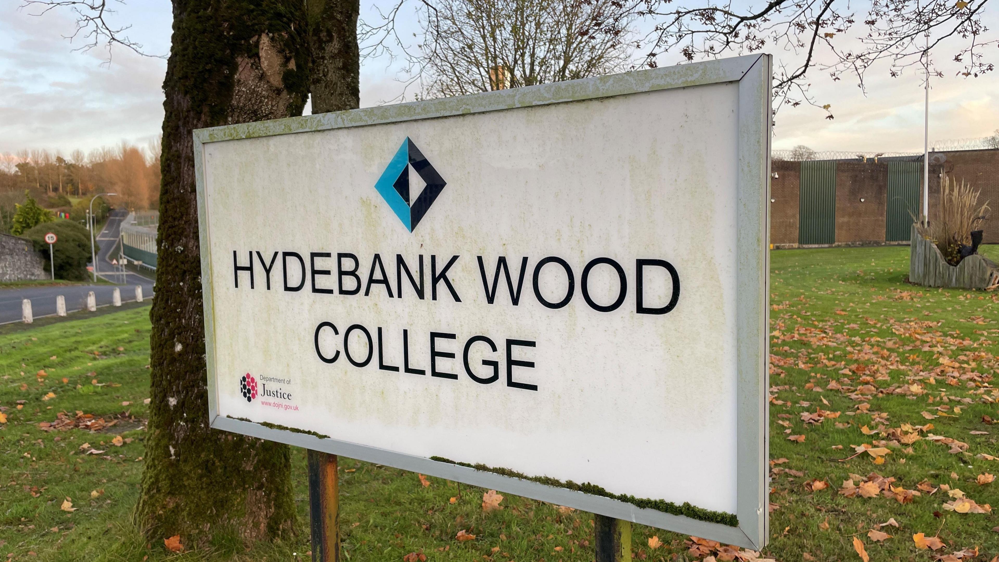 A sign saying Hydebank Wood College. In smaller writing, it says Department of Justice. A tree and leaves lying on grass are in the background.