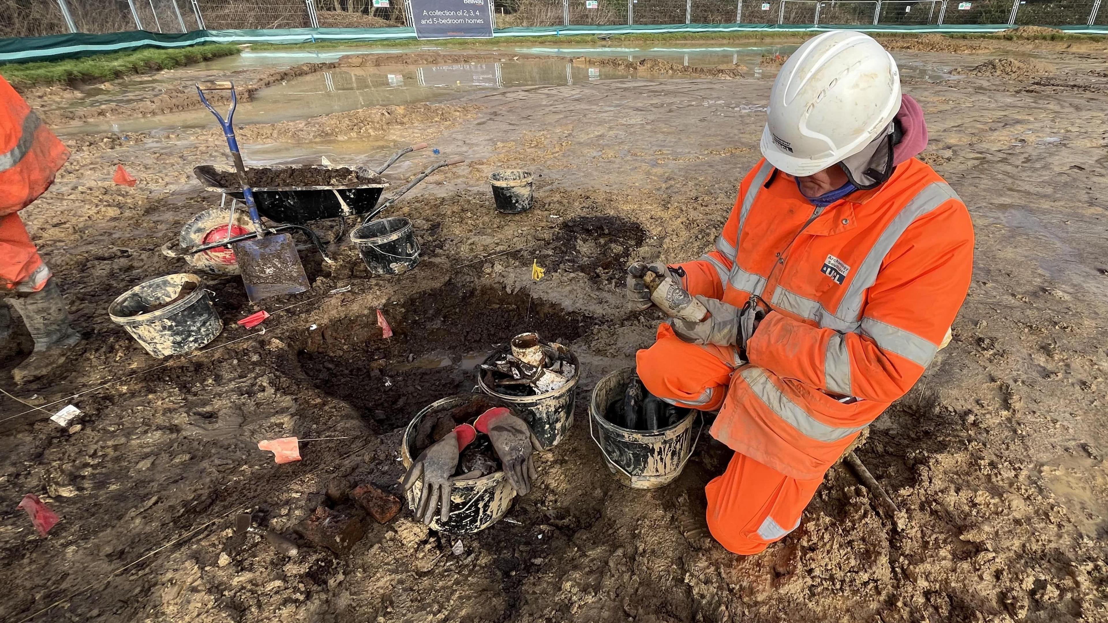 A man in a white hardhat and orange high-vis jacket and trousers on a muddy surface looking at an old glass bottle.