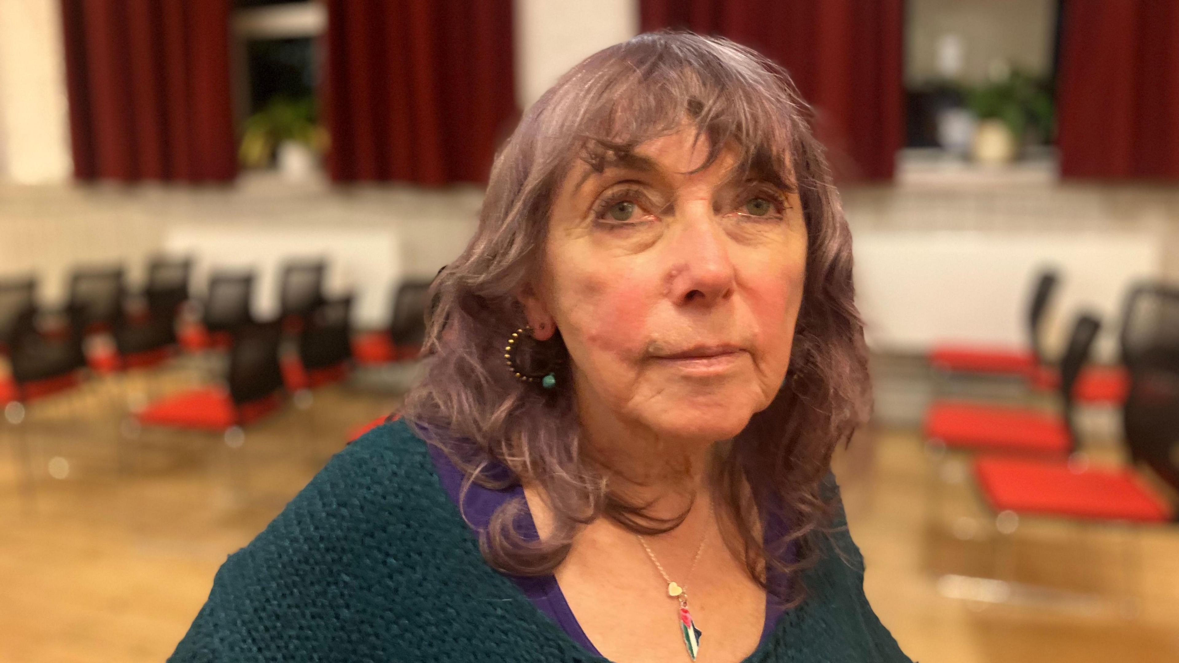 A woman with lilac grey hair wears a blue top. Behind her, a meeting space with red and black chairs is blurred.