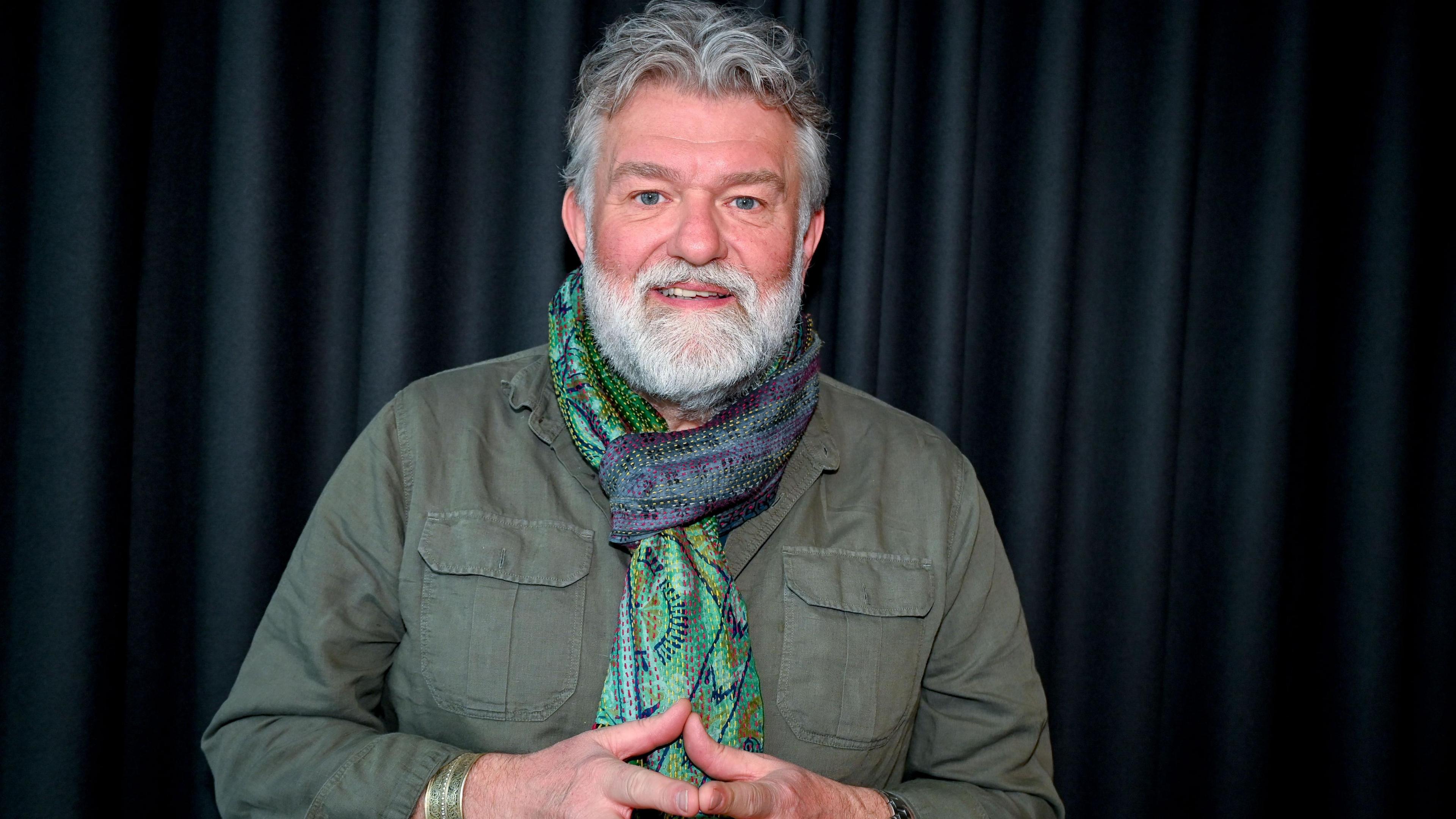Si King smiling into the camera in front of a dark, pleated curtain. He is wearing a green, blue and purple patterned scarf tied around his neck. He is wearing a green khaki shirt with large pockets on both sides of his chest. He is holding his hands together in front of his chest.