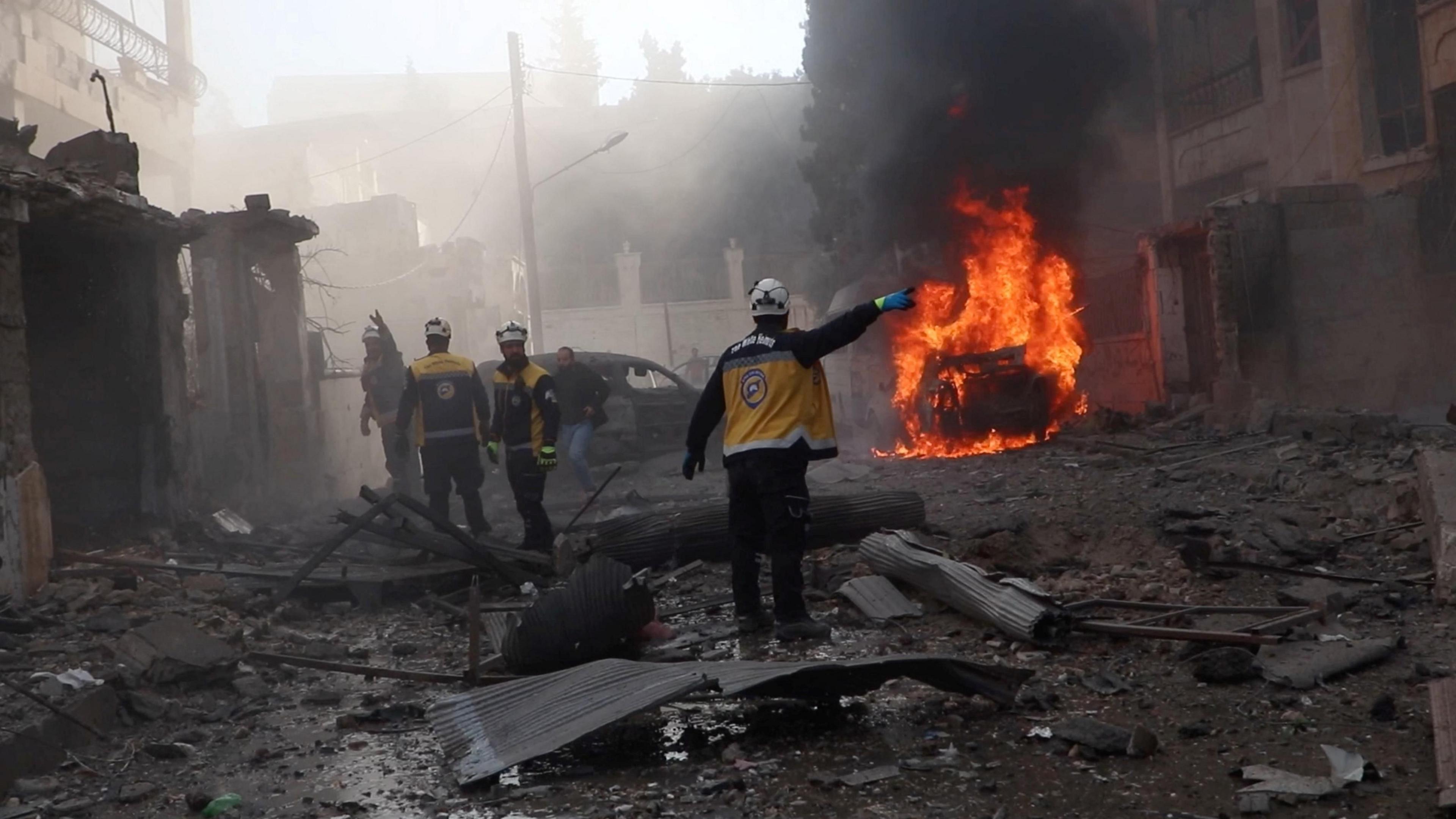 Rescue workers wearing white helmets and yellow jackets stand in a street covered in grey rubble, near a car engulfed in red flames. The air is full of smoke.