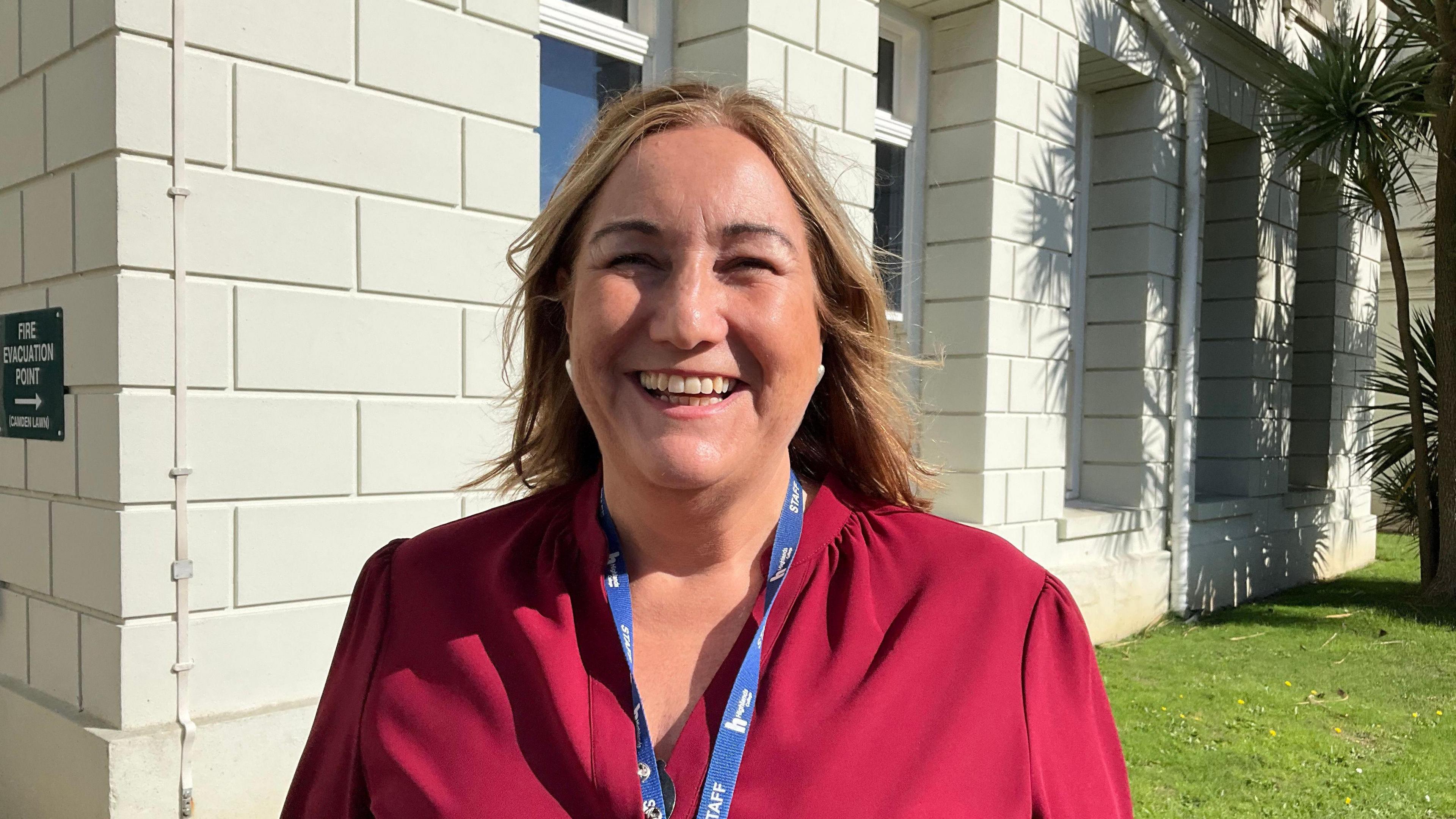 Jo Terry-Marchant is pictured wearing a dark red blouse and a blue lanyard. She is smiling widely as she stands outside a light-coloured building which is beside palm trees. A sign on the wall of the building reads 'Fire Evacuation Point'. 
