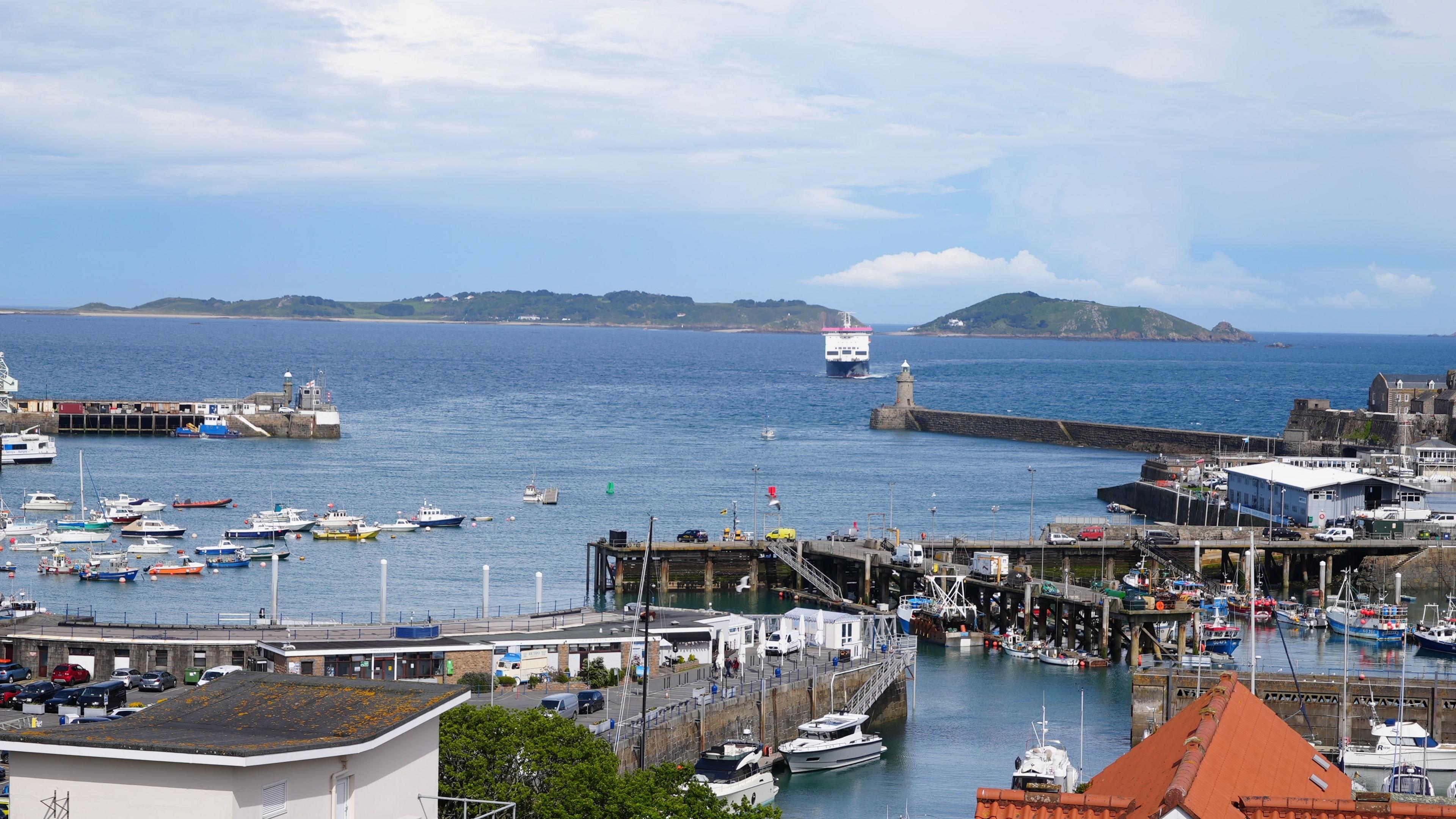 A ferry approaches St Peter Port Harbour in Guernsey. Herm and Jethou can be seen behind the ferry. Various boats, including fishing vessels, can be seen along with parts of the harbour.