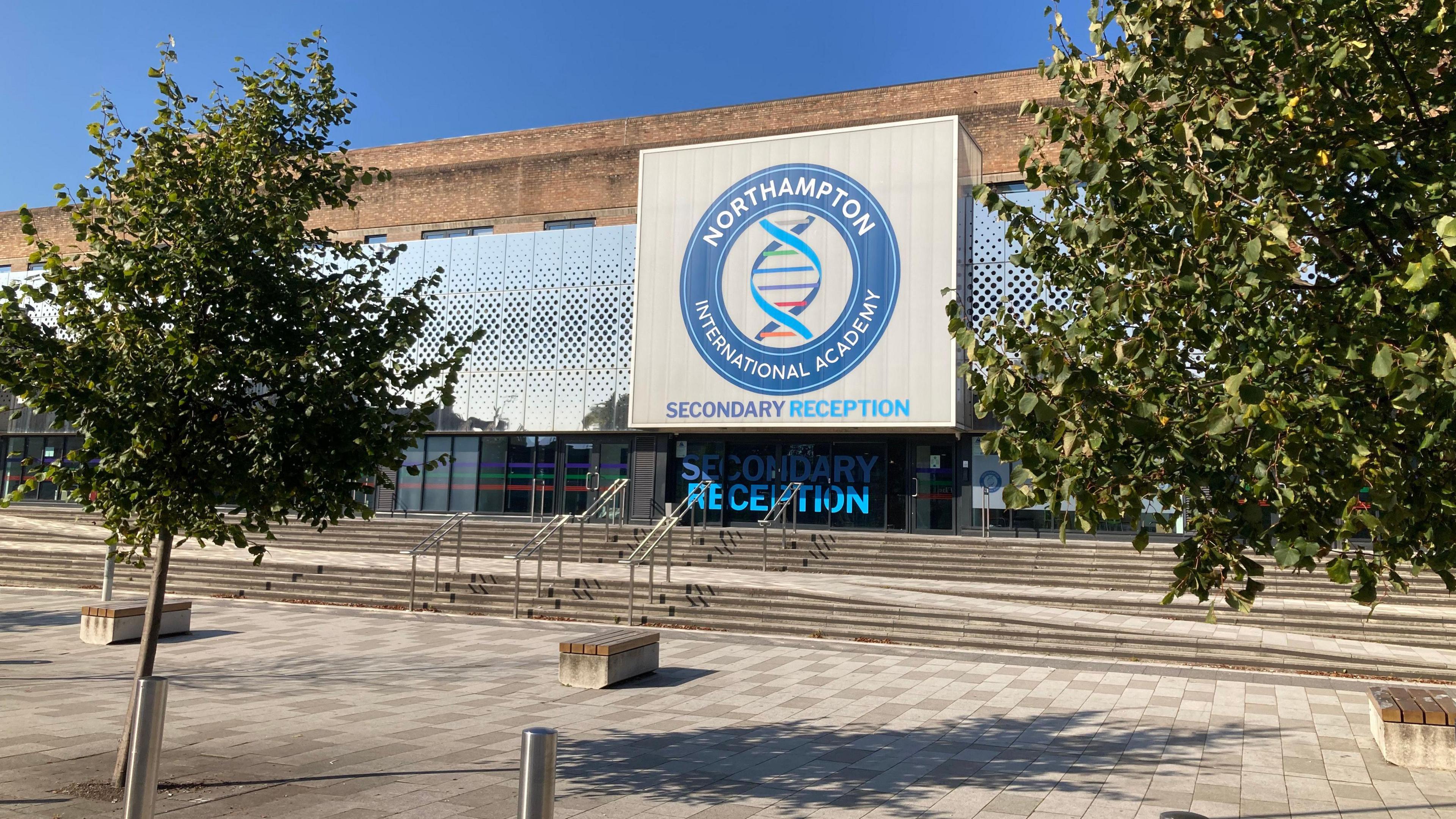 A brick building surrounded by a metal grating with a large blue square logo of the school on the front. There are lots of concrete stairs leading up to the reception. 