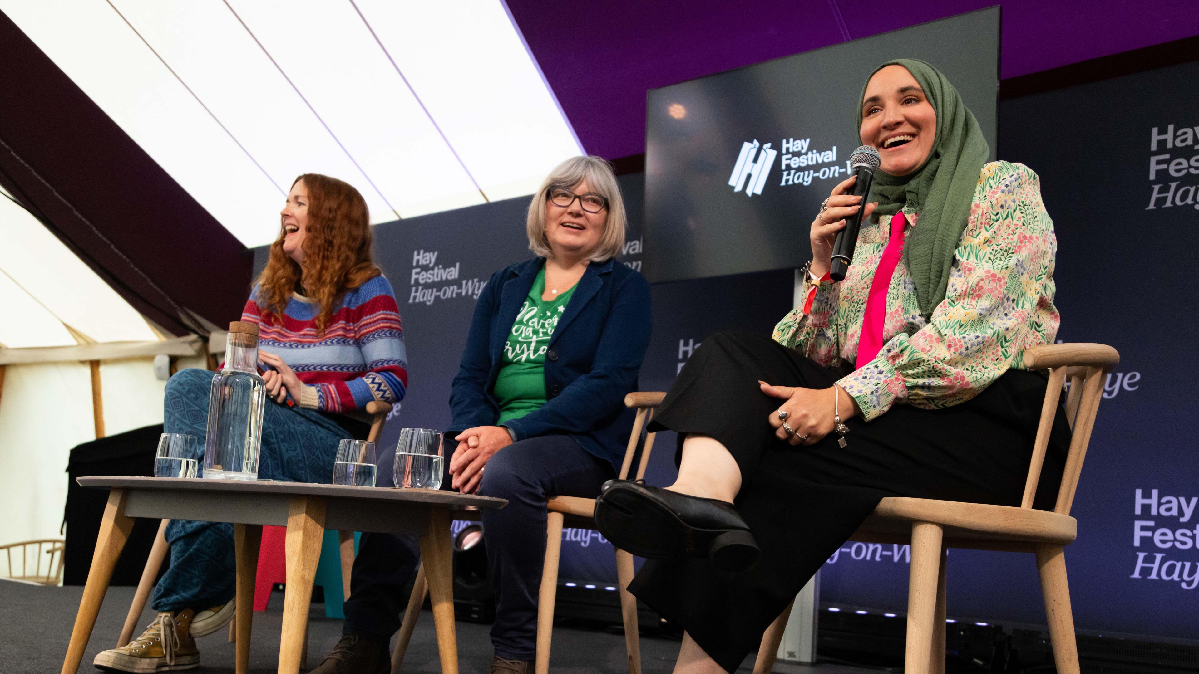 Hanan sits on a chair on a panel with two other women, a microphone in her hand. A table is in front of them with a glass jug of water and glasses. Behind them a screen has the Hay Festival logo on it.