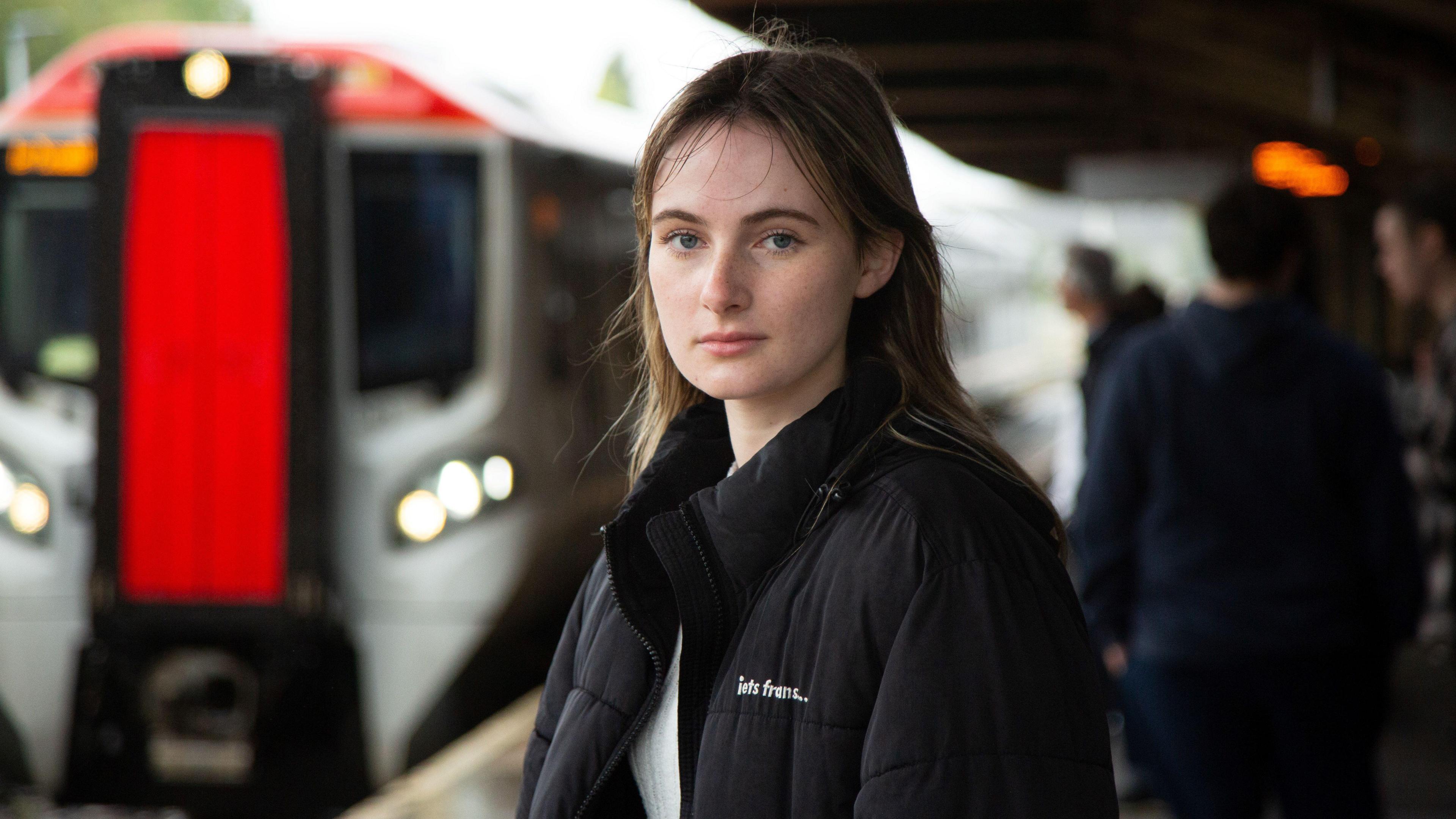 Rhiannon Williams yn sefyll ar blatfform orsaf drennau, gyda tren yn y cefndir