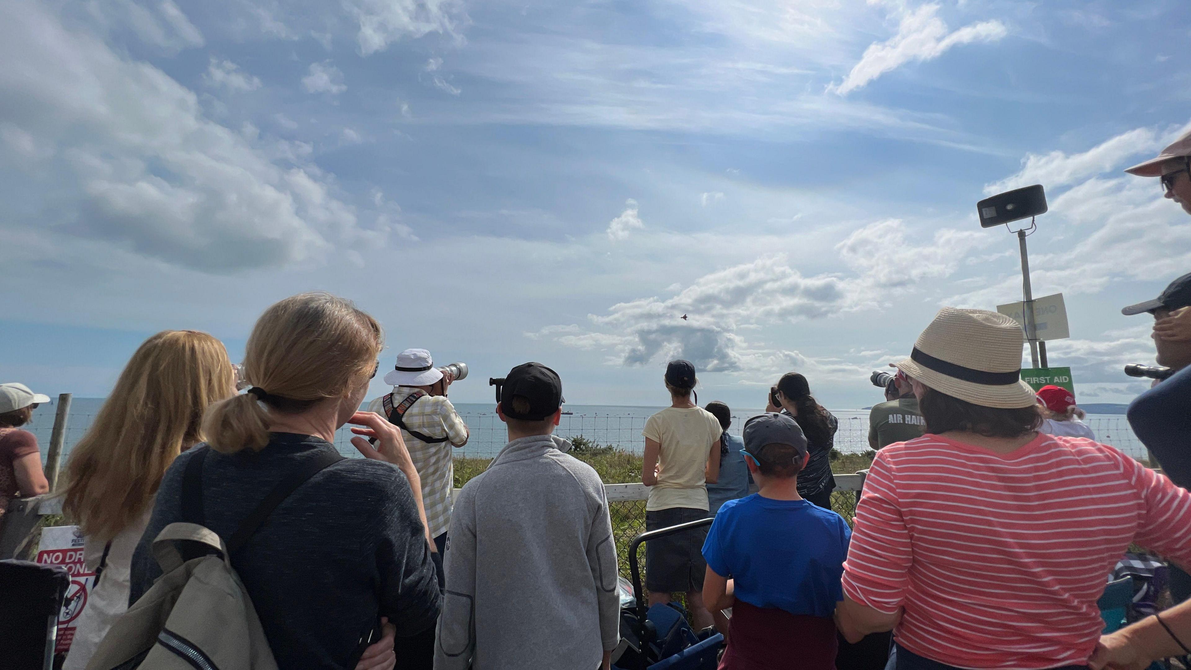 Dozens of people with their backs to the camera looking out to sea, where in the sky there are planes flying.