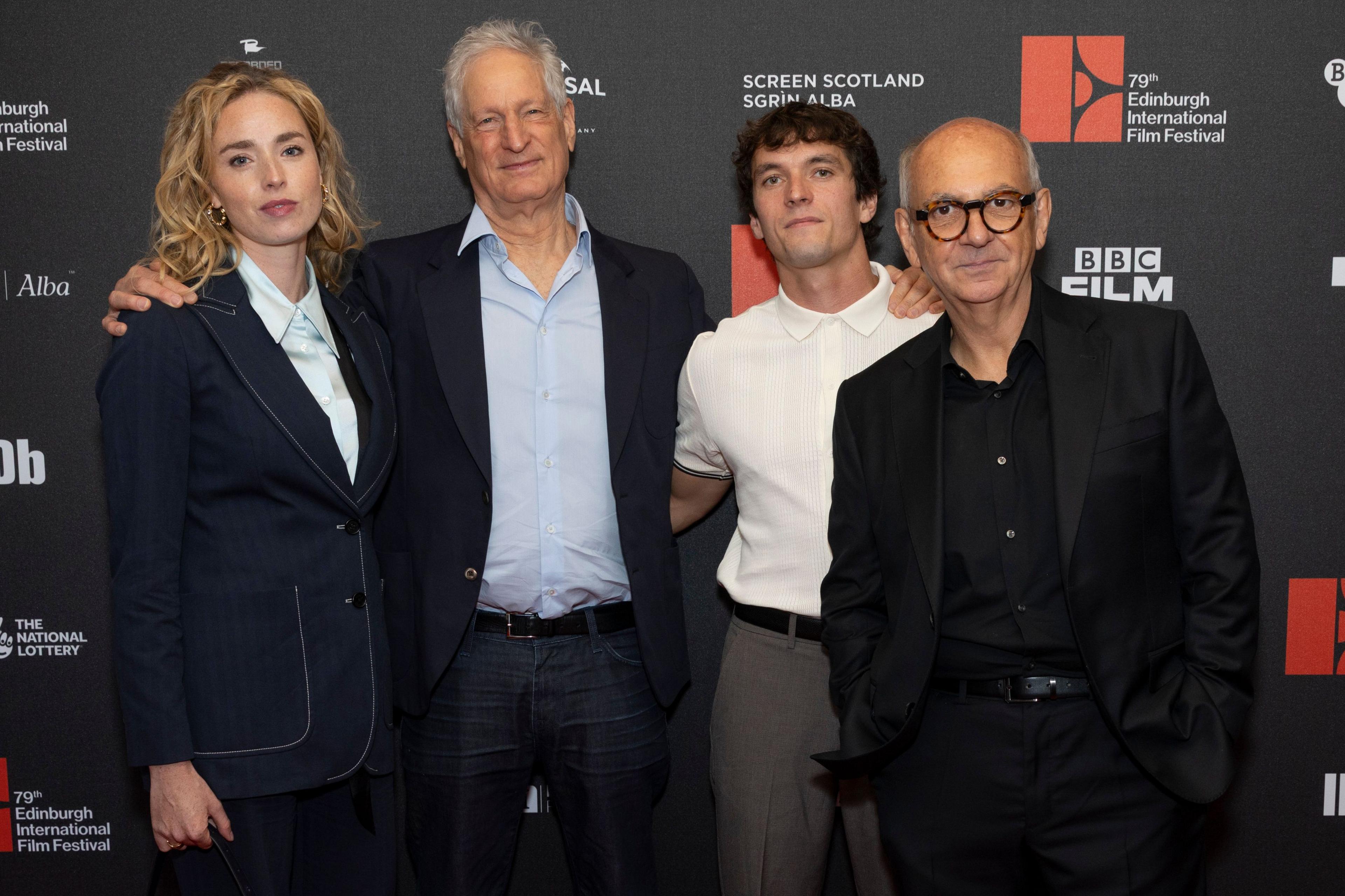 Freya Mavor is in a blue suit and white shirt and has long blonde hair. Marc Turtletaub has white hair and is in a blue blazer, grey trousers and light blue open-necked shirt. Fionn Whitehead sports a white short-sleeved shirt buttoned at the neck and Luis Gnecco is in a black suit and shirt. They stand in a row, arms around one another, in front of a black, white and red Edinburgh International Film Festival board.