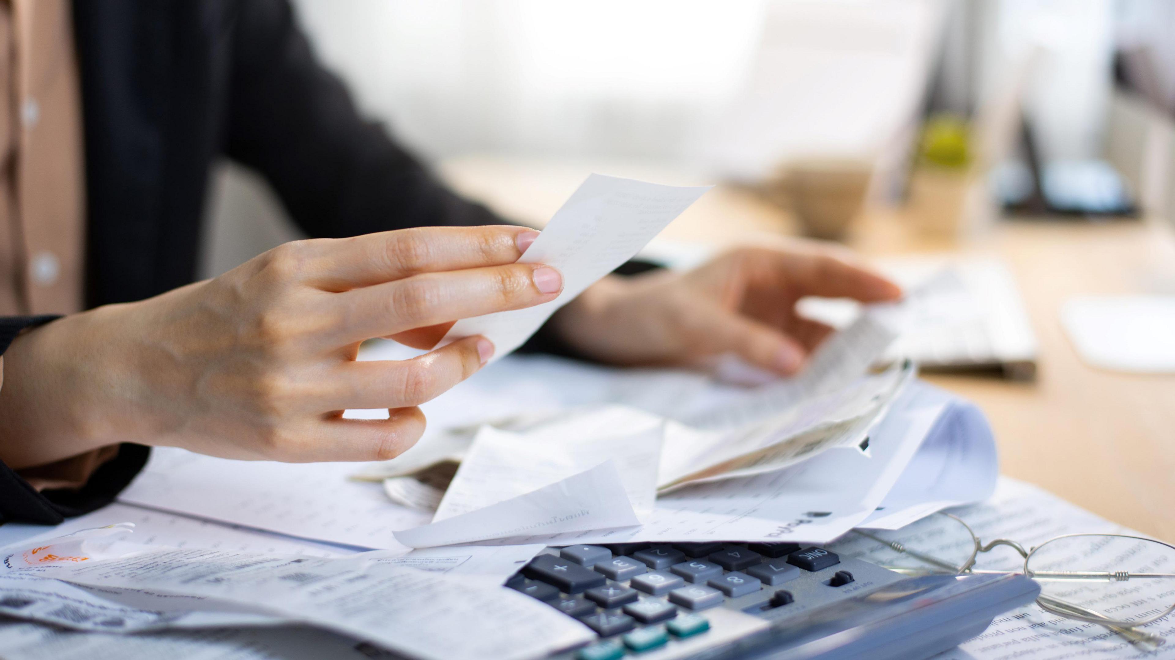 Hands holding up receipts, with a calculator and glasses on the table.