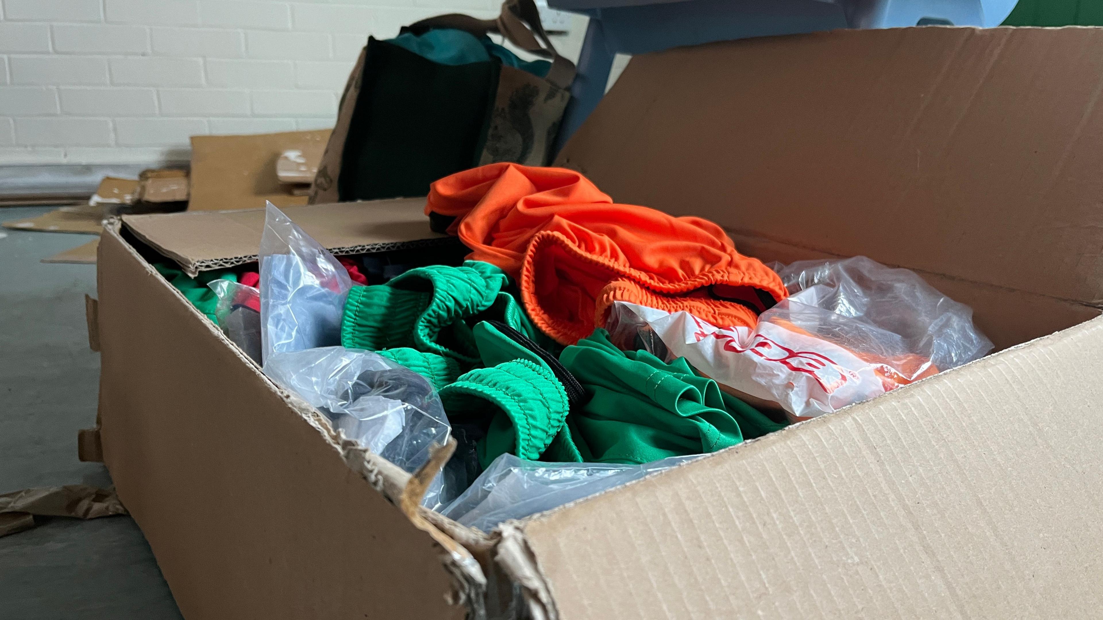 A brown cardboard box on the floor. It is filled with bright coloured clothing. 