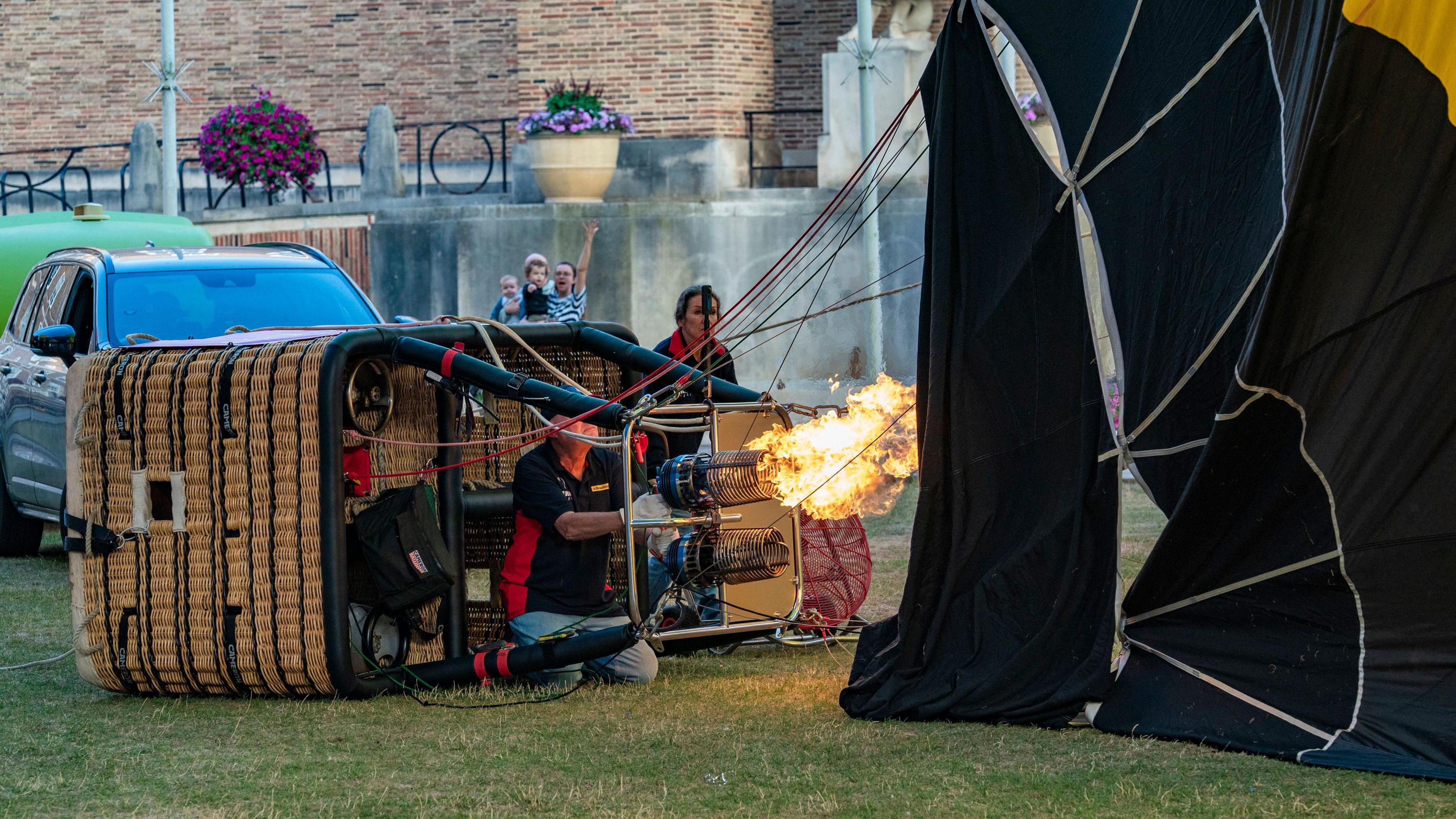 A man and a woman tending to a hot air balloon. The man is sat by the basket, which is on its side, as flames shoot out of the burner into an inflating balloon canopy. The woman is behind stood behind the basket operating a large fan. 