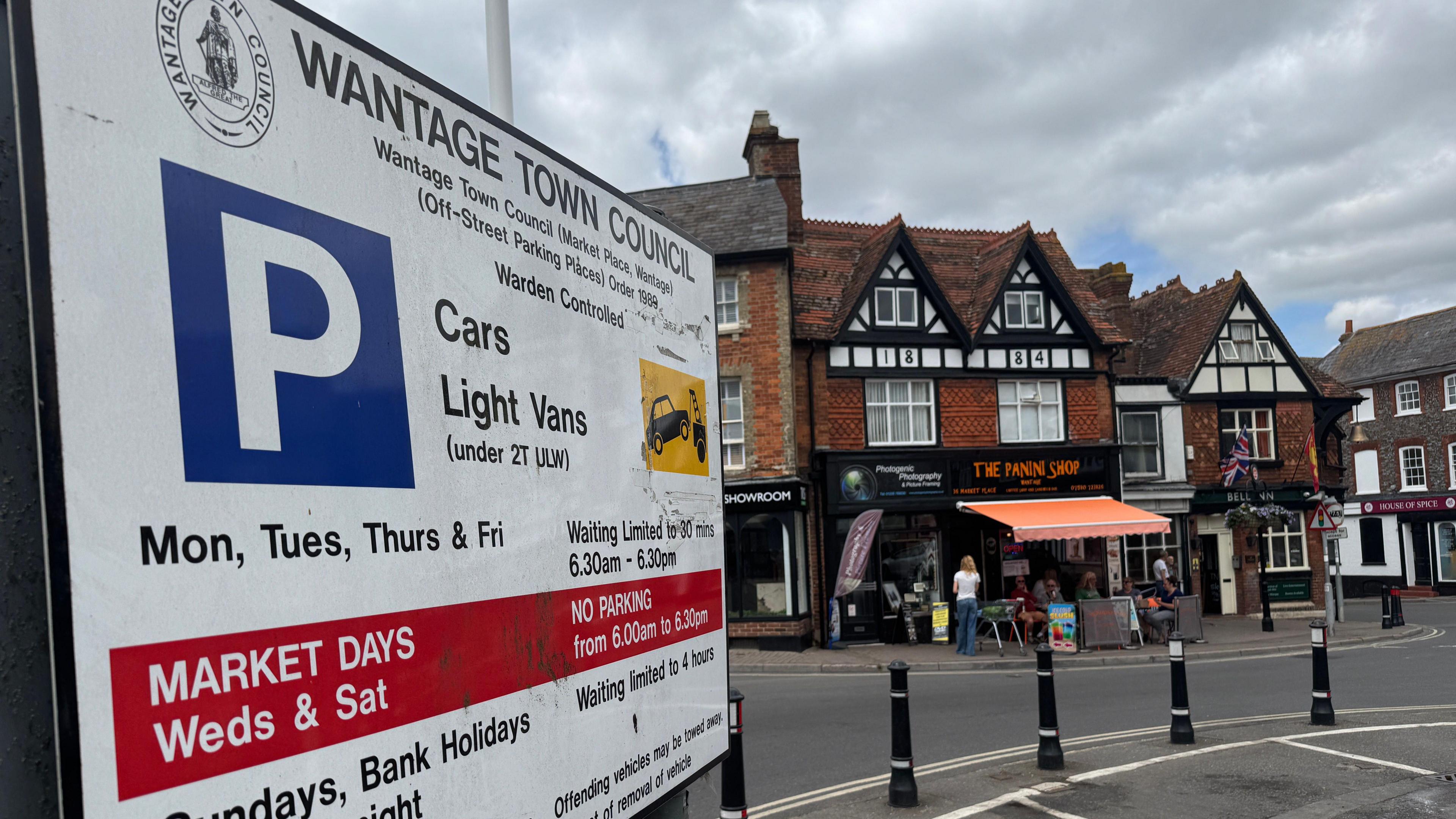 Wantage's Market Place. A small car park is pictured at the centre of a large town square. On the left is a parking information sign which gives information on parking times and costs. In the background are high street shops with a cafe outside featuring outside seating