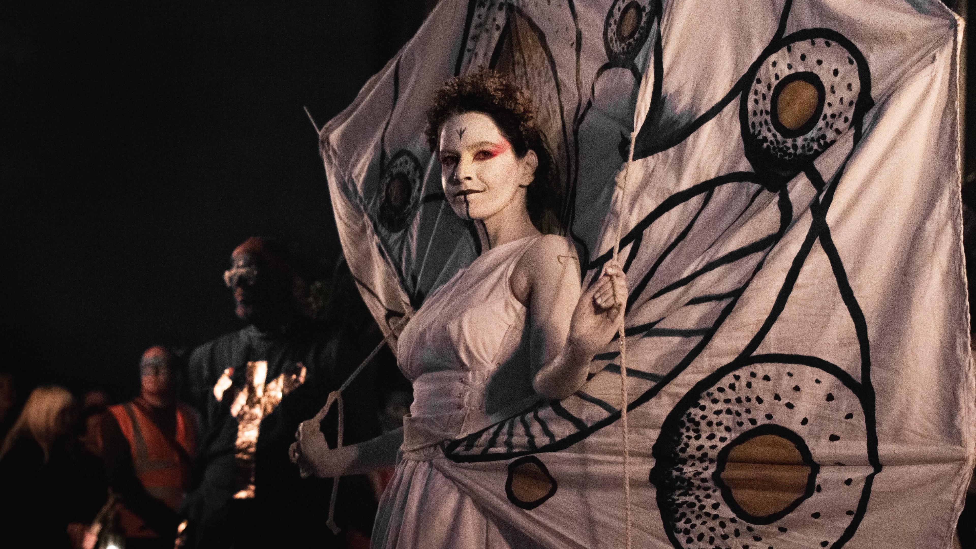The May Queen wearing all white and white face paint with a large white banner behind her.