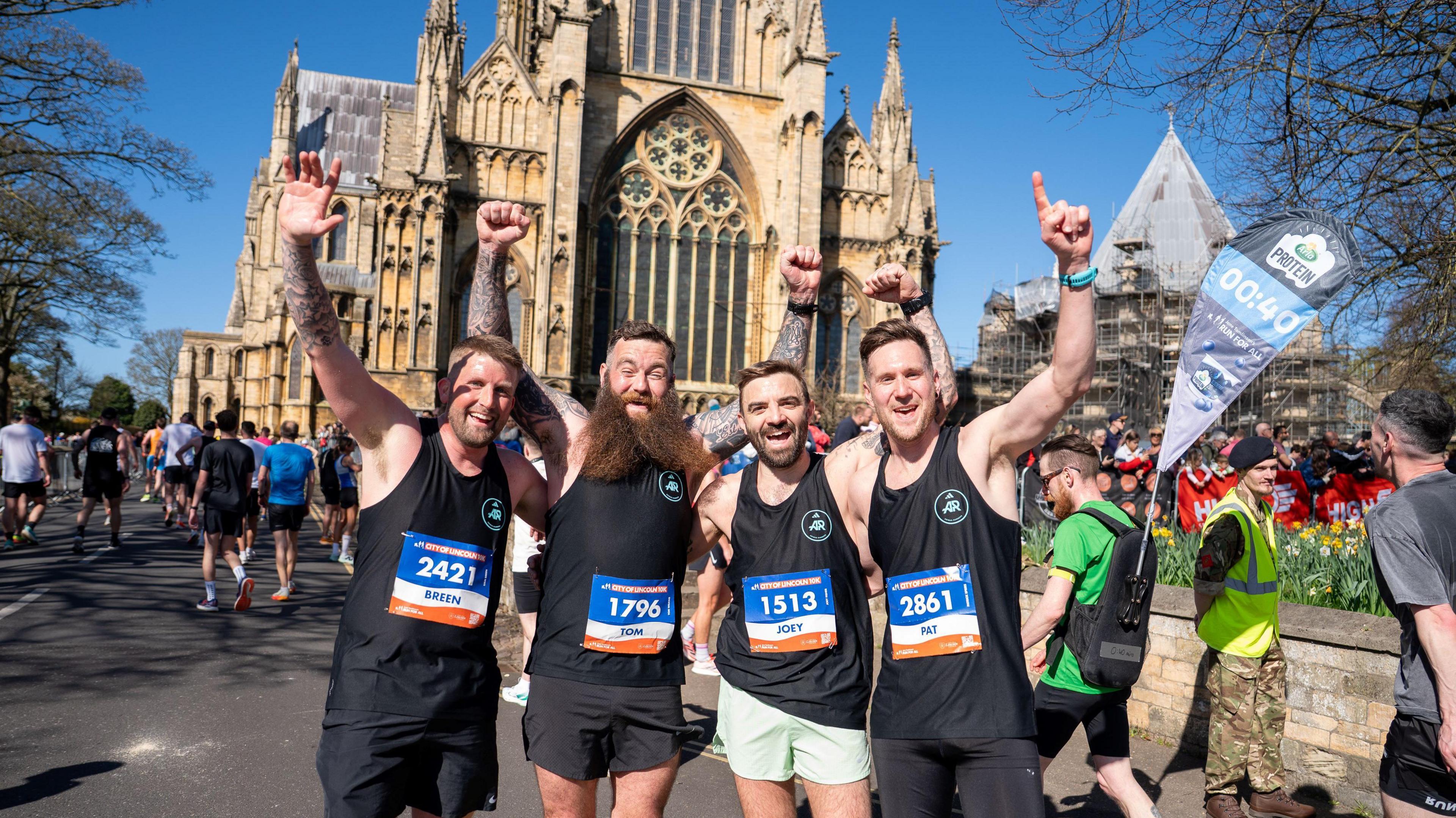 Four male runners in black sportswear waving to the camera in front of the Lincoln Cathedral. There are other runners in the background and a man in a hi-vis vest and a hat.