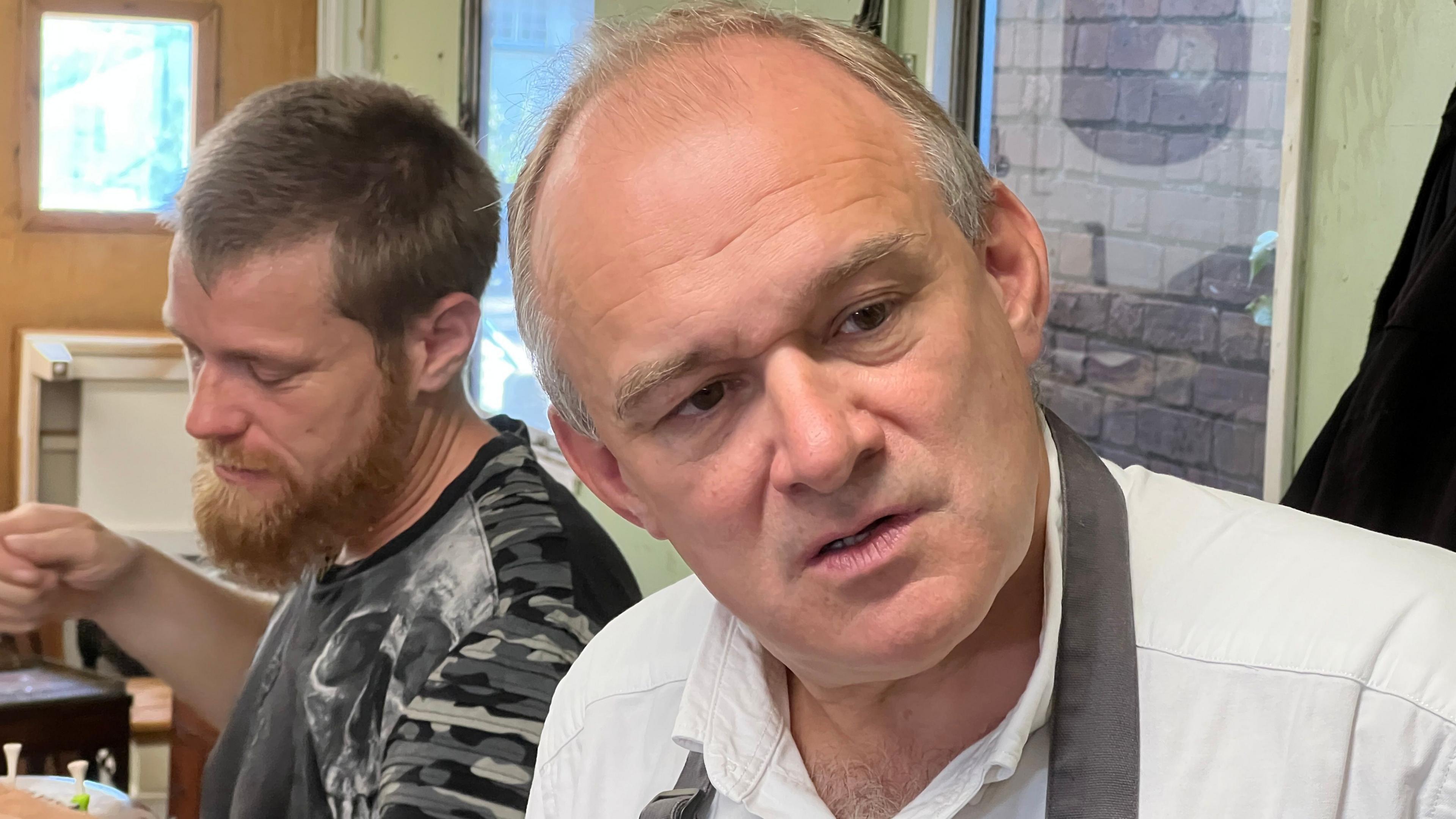 Ed Davey listening looking concerned,  behind him is a man with a beard working.