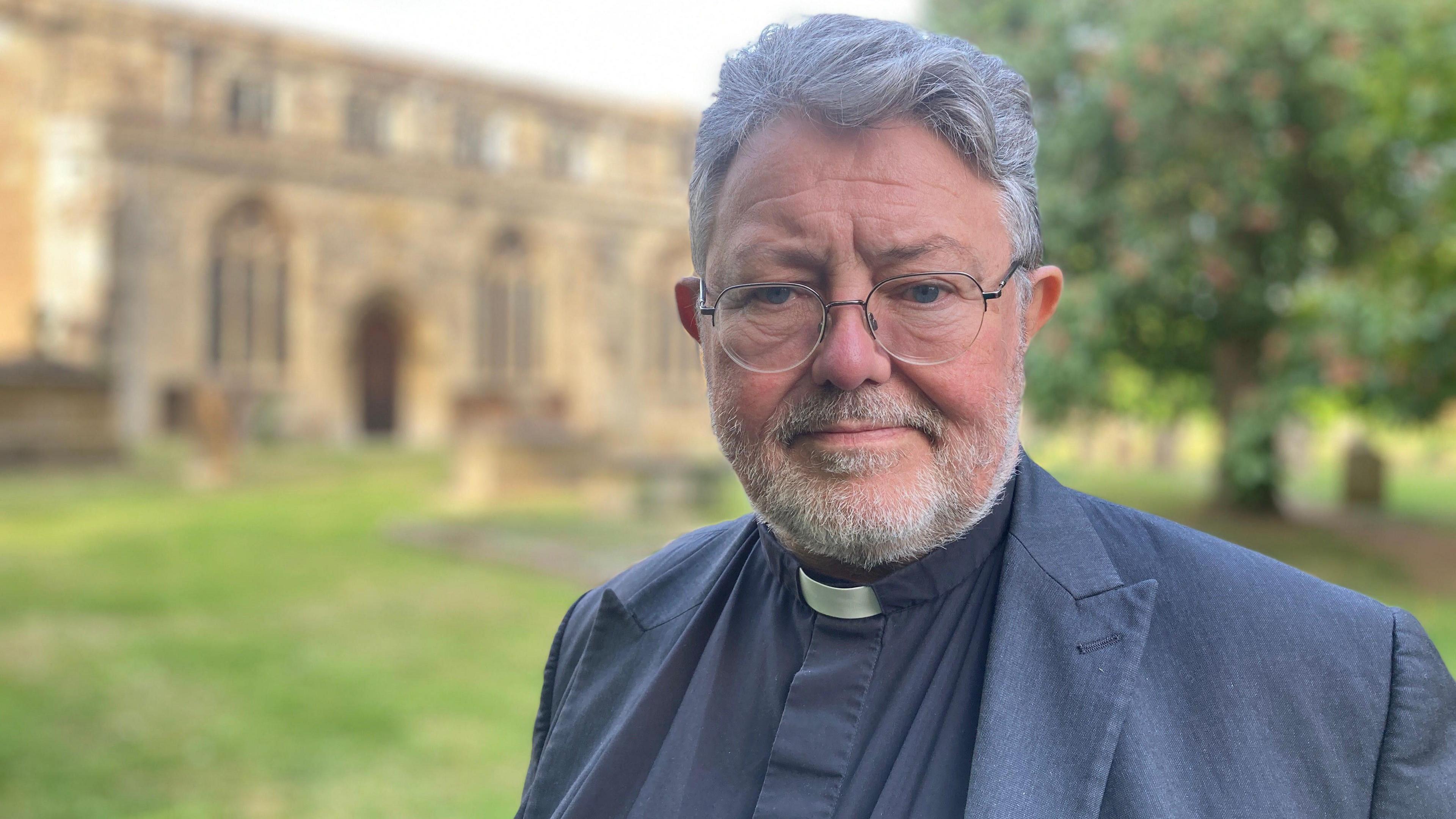 A man with grey hair and stubble, wearing glasses, a black suit and a clerical collar. He's looking at something off-camera, and standing in a graveyard with a church behind him.
