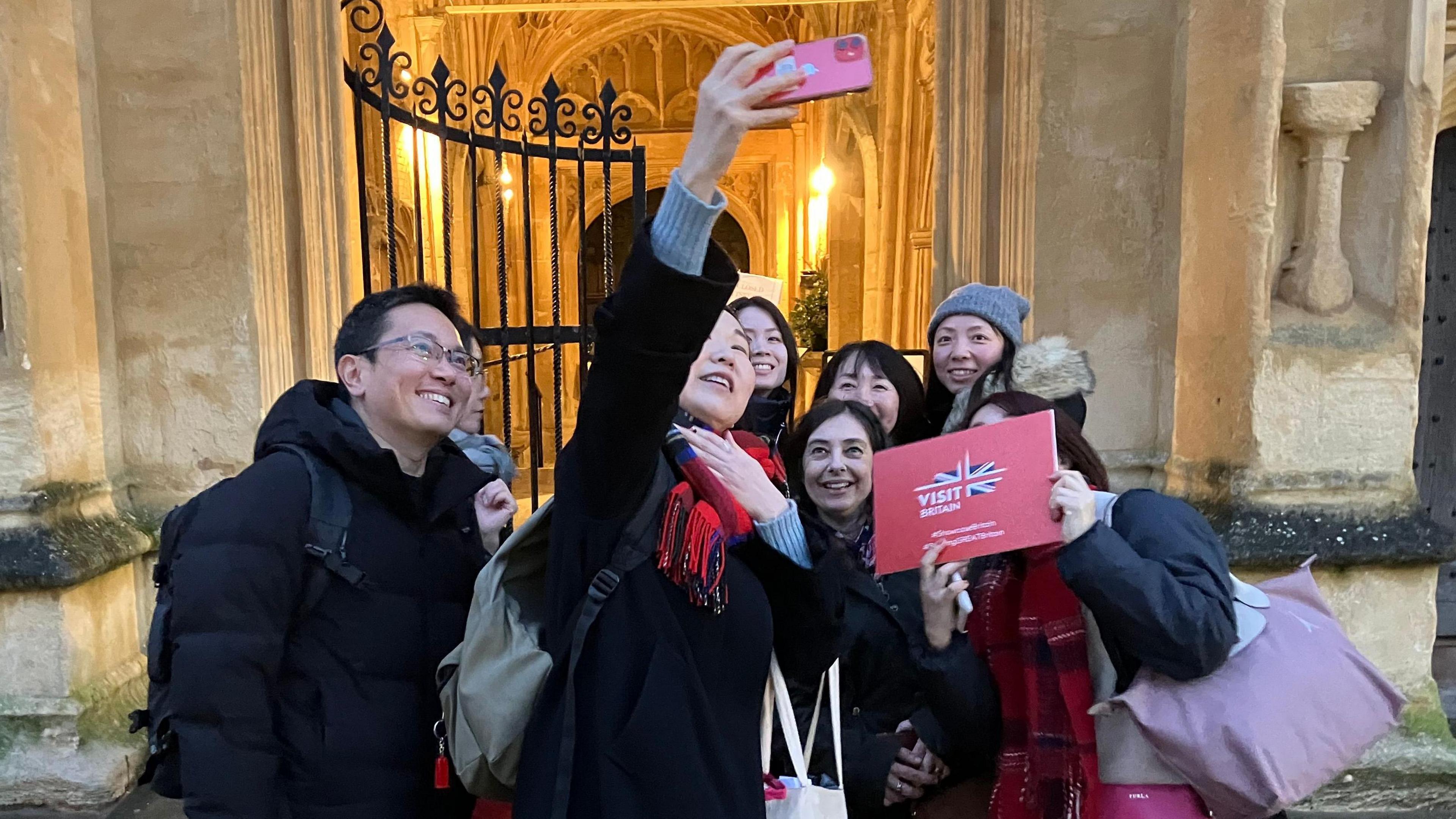 A group of Japanese tour operators huddles together as a woman in the middle takes a selfie on a pink phone. Everyone is wearing a coat and they are standing outside a cream-coloured old stone building.
