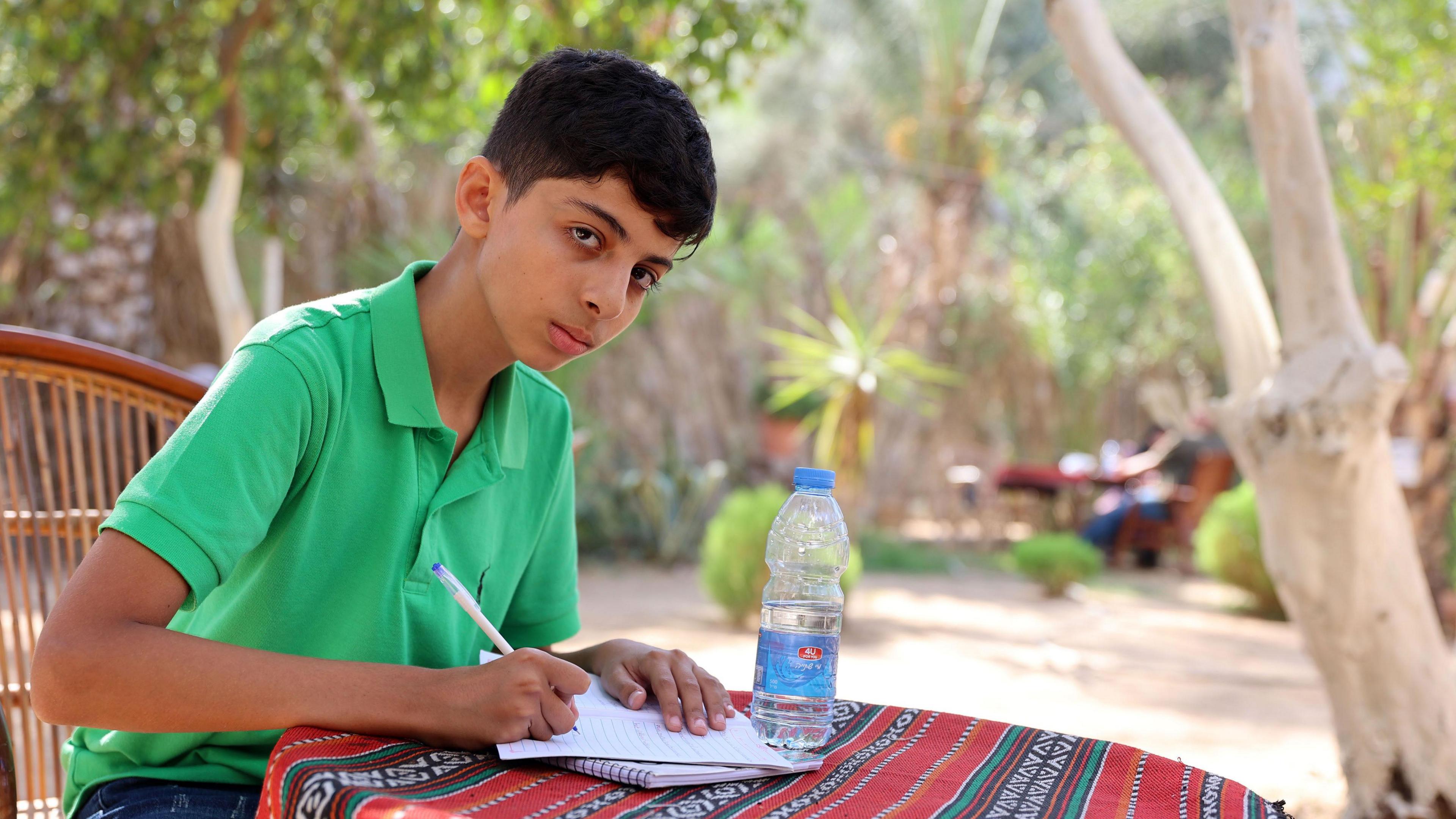 Abdullah wears a green polo shirt as does his schoolwork on a table in a garden. He is writing on paper with a pen as he looks up towards the camera.