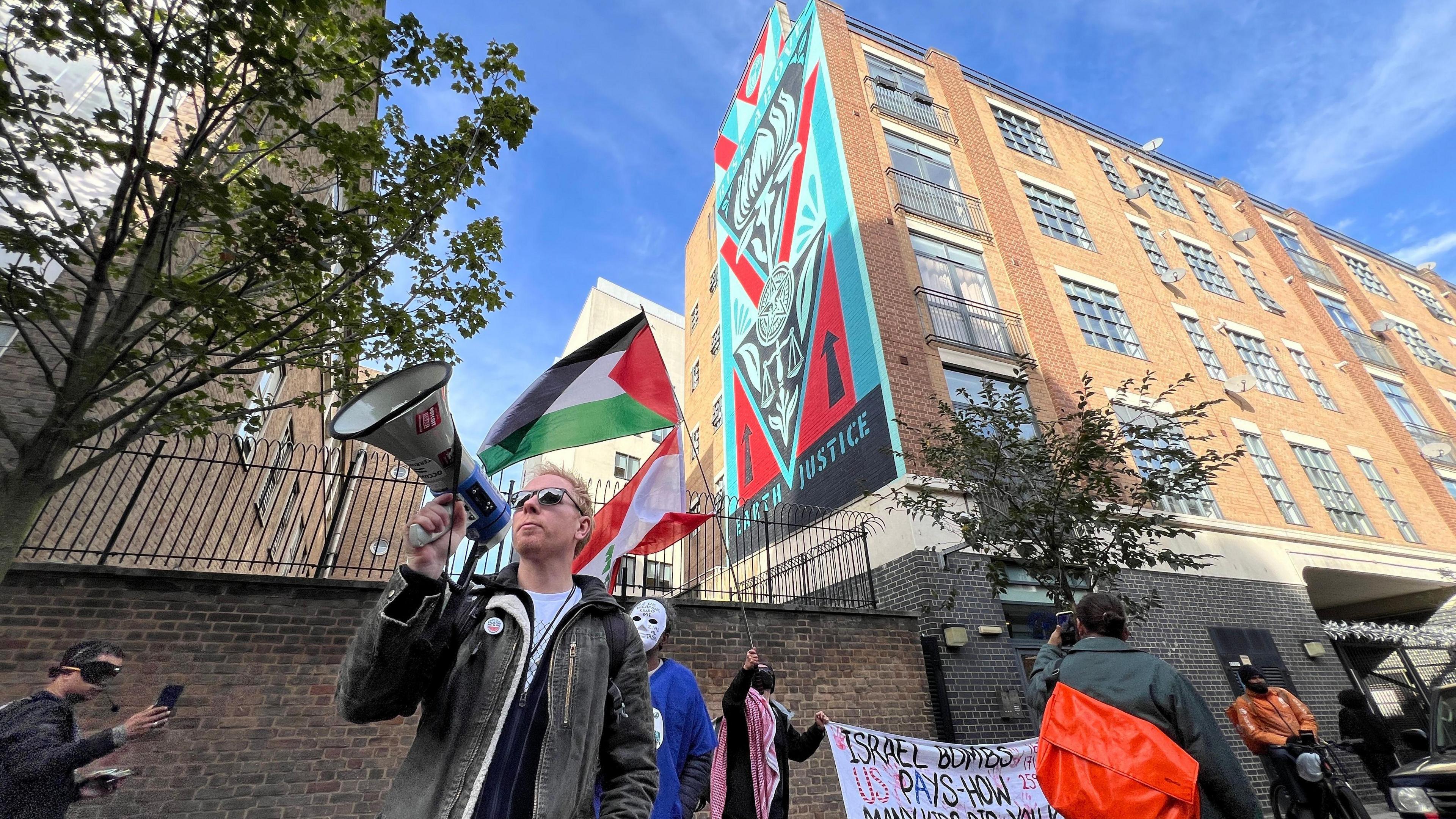 A man with a megaphone stands in front of a four storey high mural while protestors wave a Palestinian flag in the background