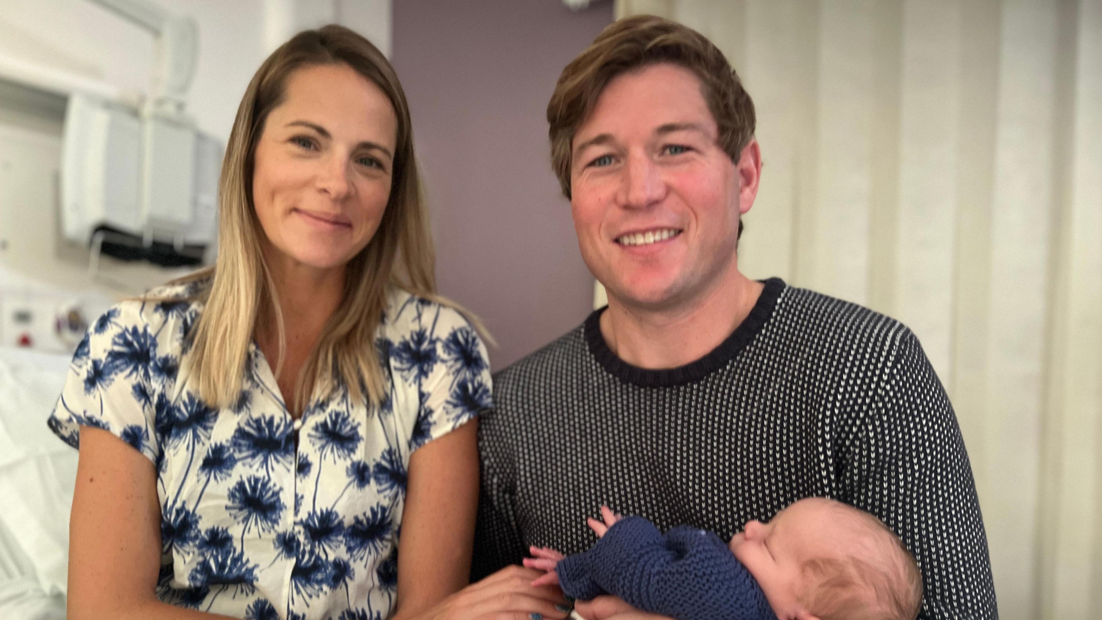 Jessica Williams with below shoulder-length fair hair, wearing a white top with large blue flowers. Sitting beside her is her husband Pete, with short light brown hair, wearing a black and white jumper. Both are smiling towards the camera. He is holding their son Orlando, who is sleeping and wrapped in a dark blue knitted cardigan.