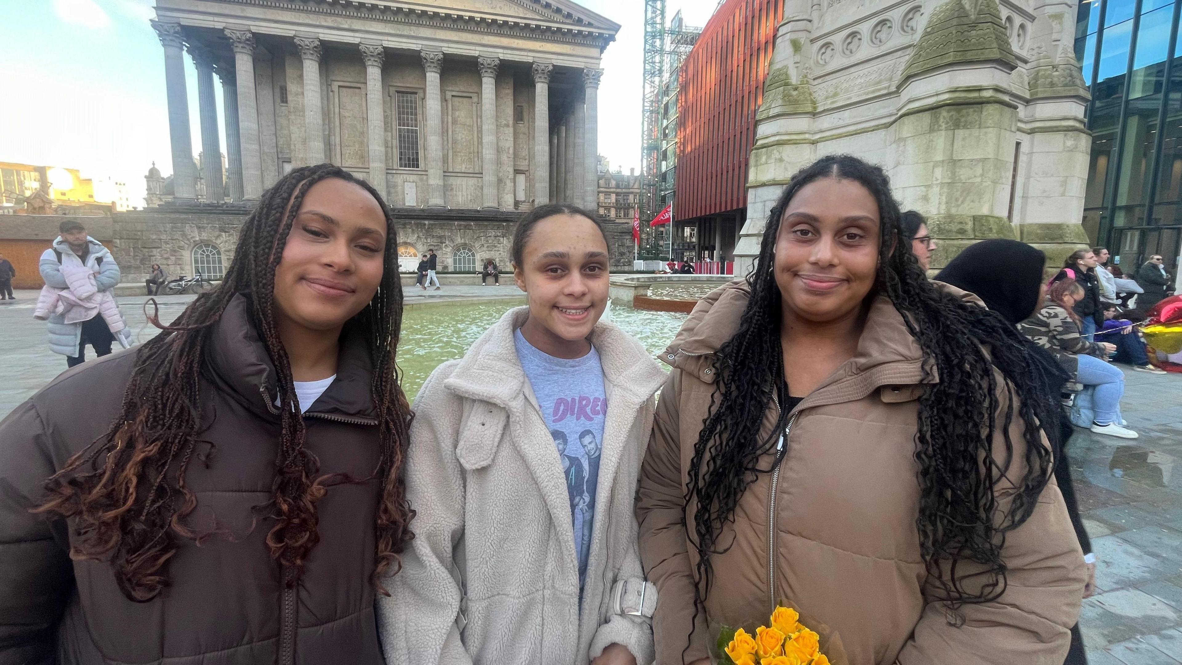 Three young women stand together smiling at the camera, in front of a fountain and a building with pillars. The two women on the sides wear brown coats, while the young woman in the middle wears a white coat, with a grey One Direction t-shirt underneath. The girl on the right is carrying a yellow bouqet of flowers.