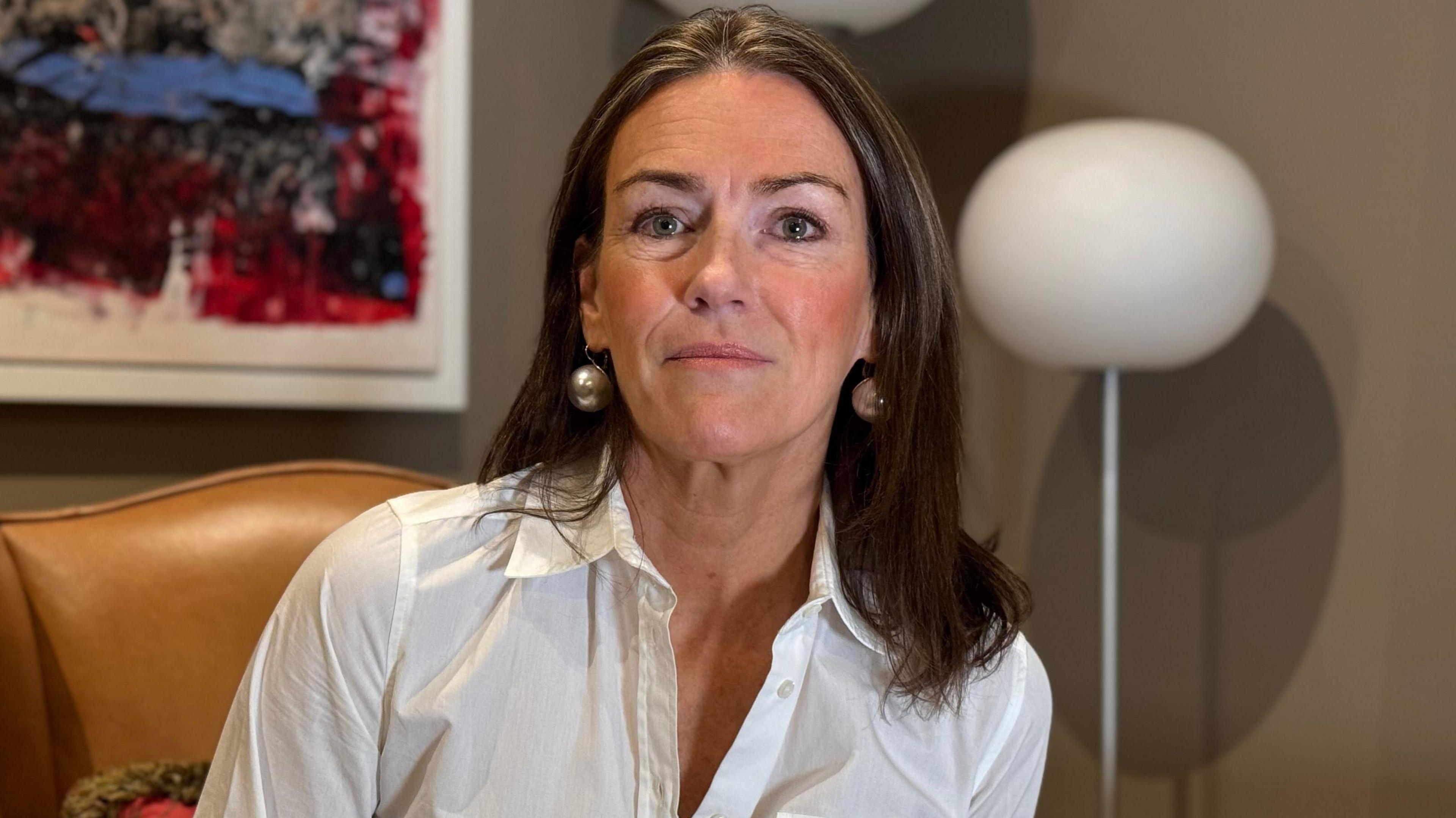 Helen O'Sullivan sitting inside on a brown leather chair. There is a painting and lamp behind her. She is wearing a white shirt, silver earrings and has brown hair. 