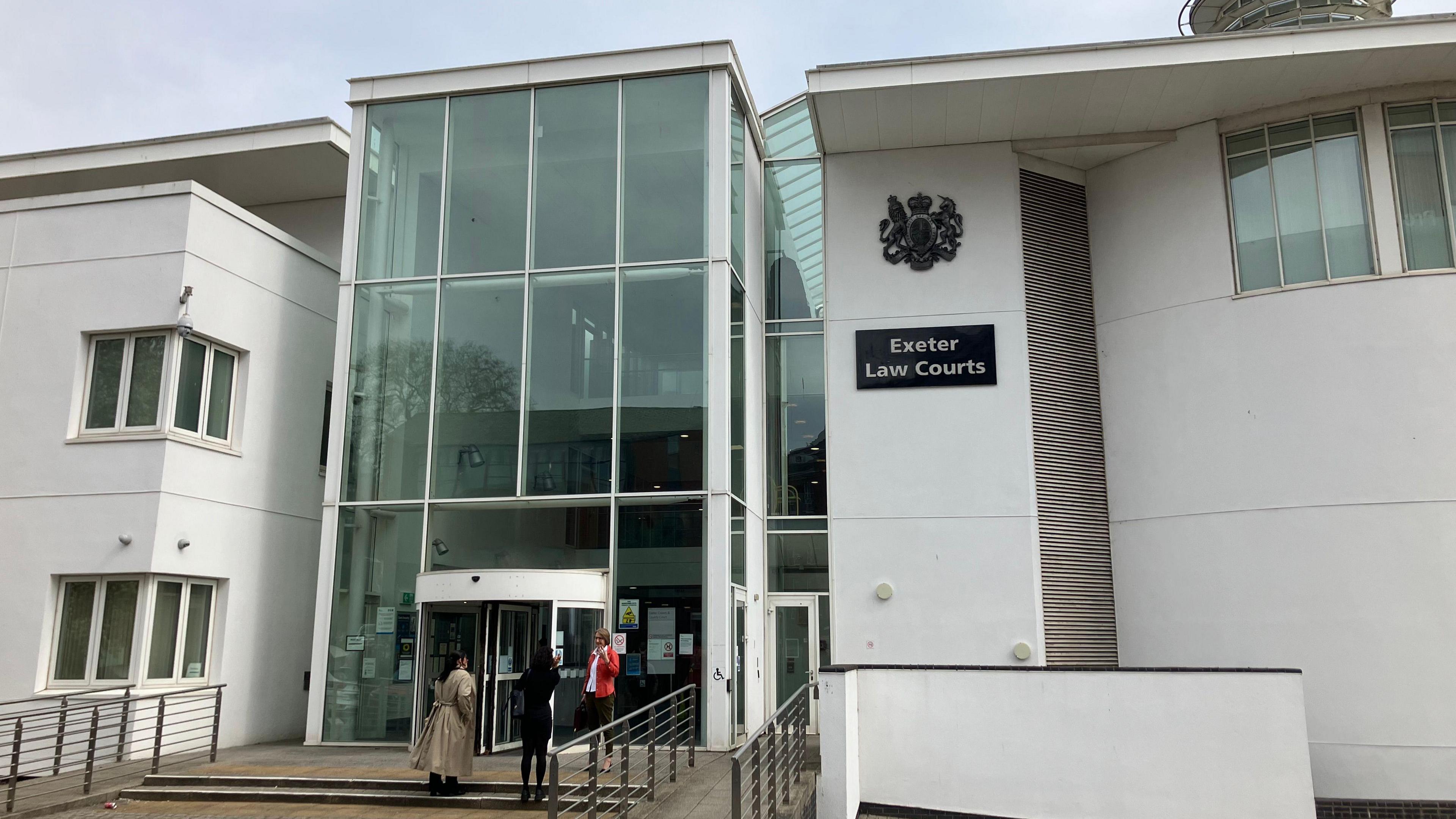 A general view of Exeter Law Courts. It is a white building with a glass paned section in the middle. Three people are standing outside.