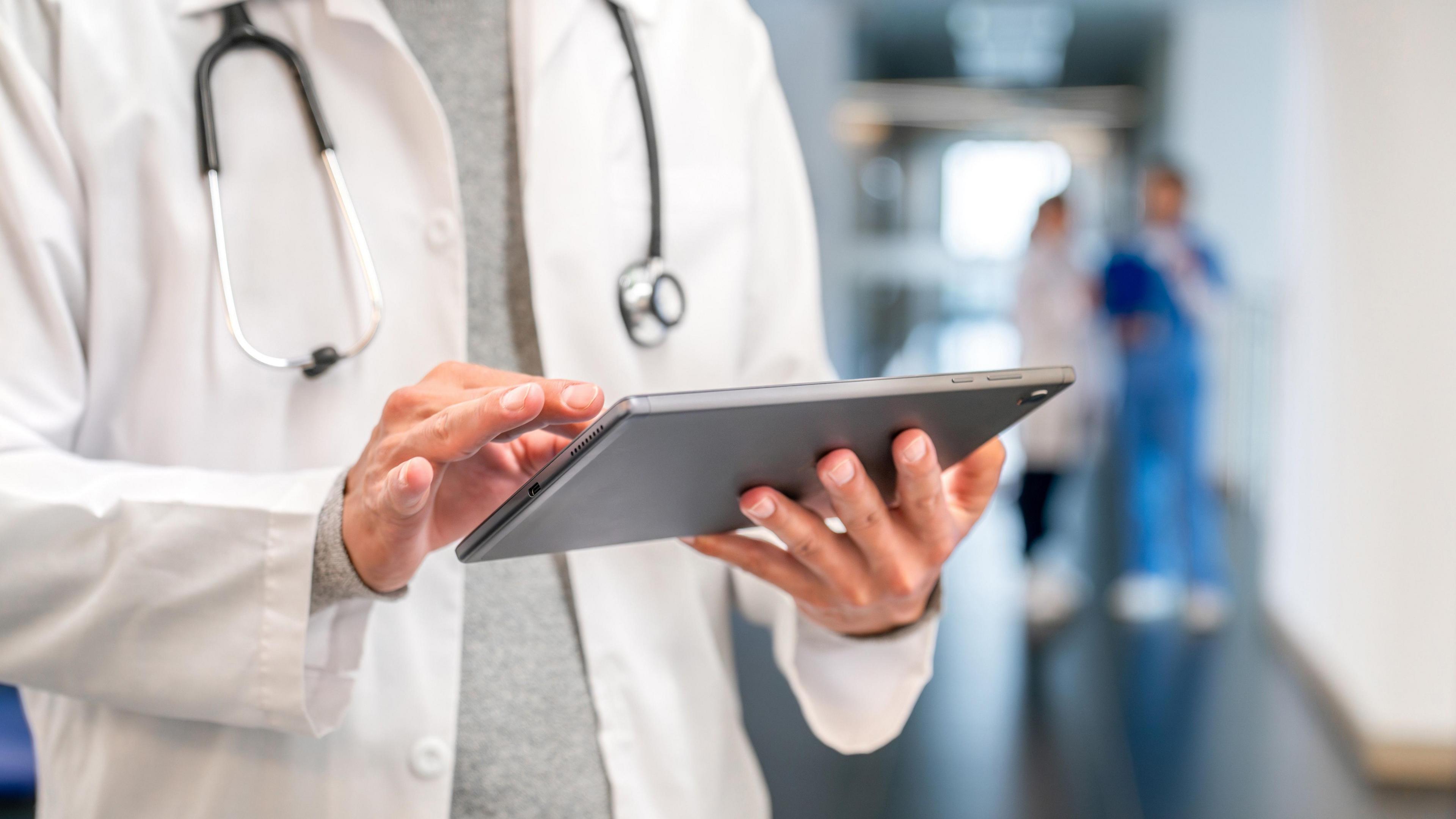 Close up of a doctor wearing a white coat and standing in a hospital corridor, looking at the screen of a tablet.
