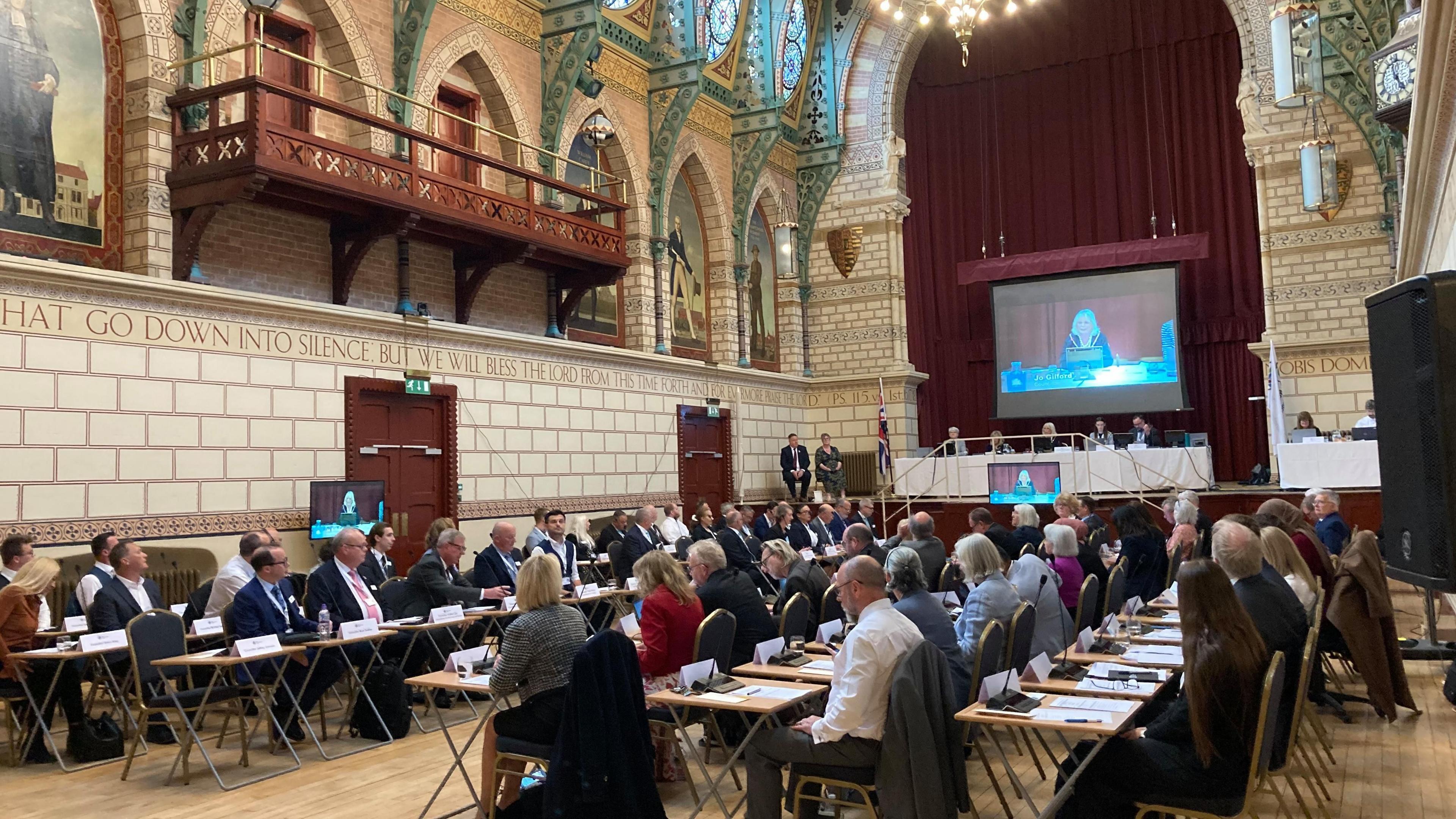 West Northamptonshire Councillors sat at desks with microphones with several people sat up on a stage