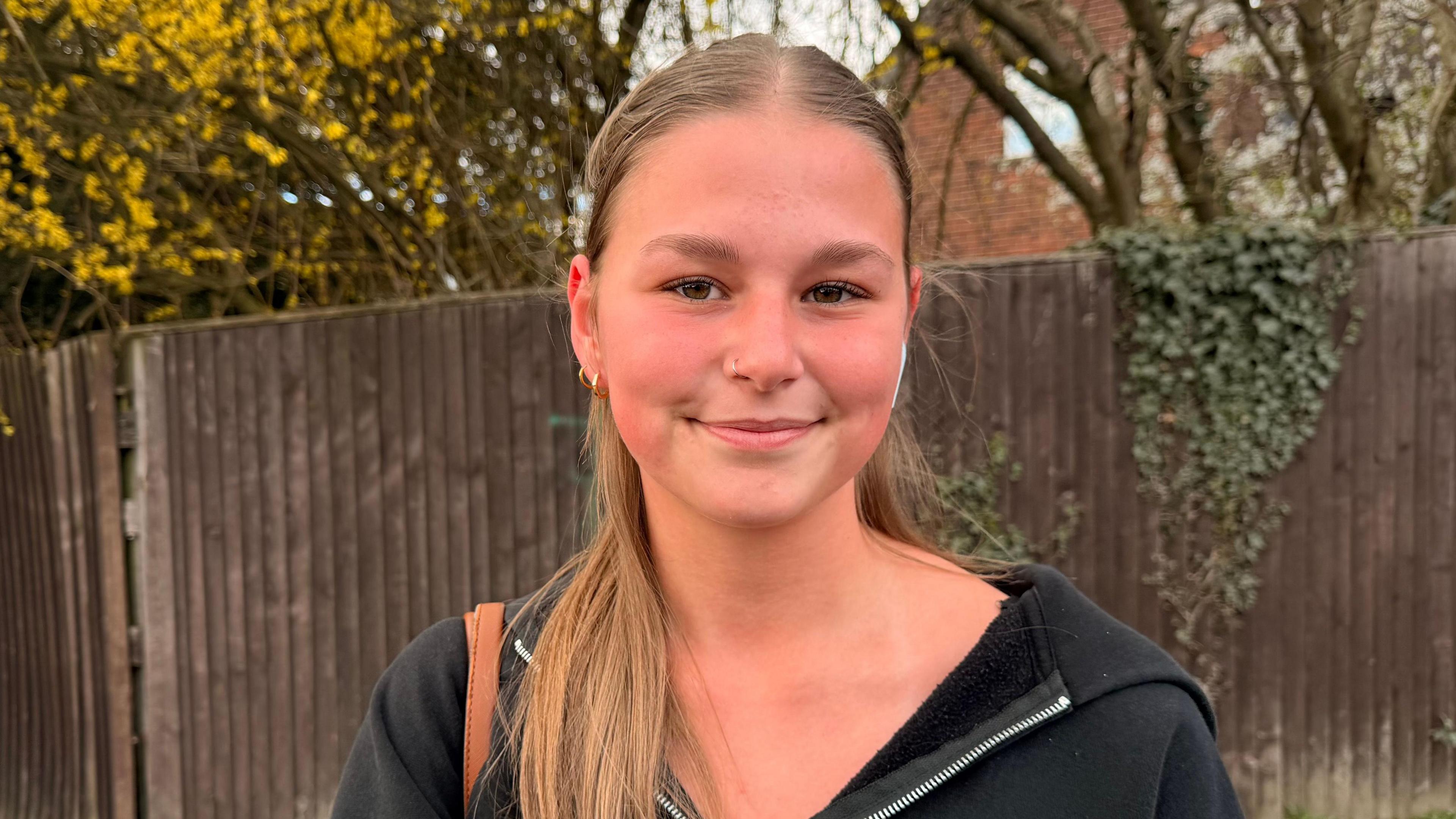 A young woman with light brown hair and brown eyes smiles as she stands in front of a brown wooden fence and trees. She wears a black zip-up jumper and wireless, in-ear headphones. 