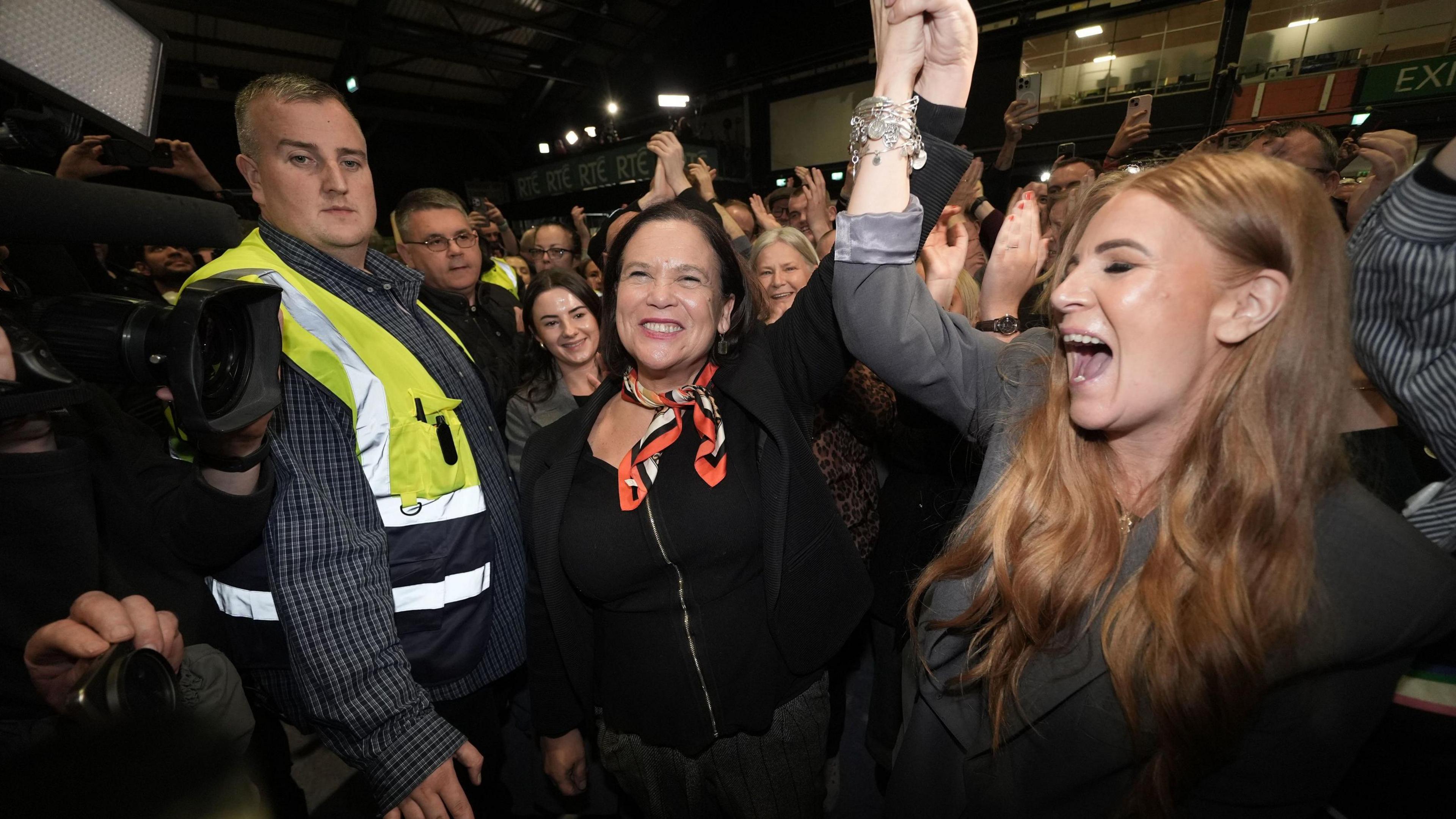 Mary Lou McDonald smiling with her arm raised in the air as she celebrates, dressed in a black top and patterened necktie... A younger woman with light brown hair, dressed in a grey blazer, is cheering and holding Mary's left hand up. To her right is a man in a checked grey shirt and hi-viz yellow waistcoat, there are press cameras visible and a large crowd of smiling supporters in the background