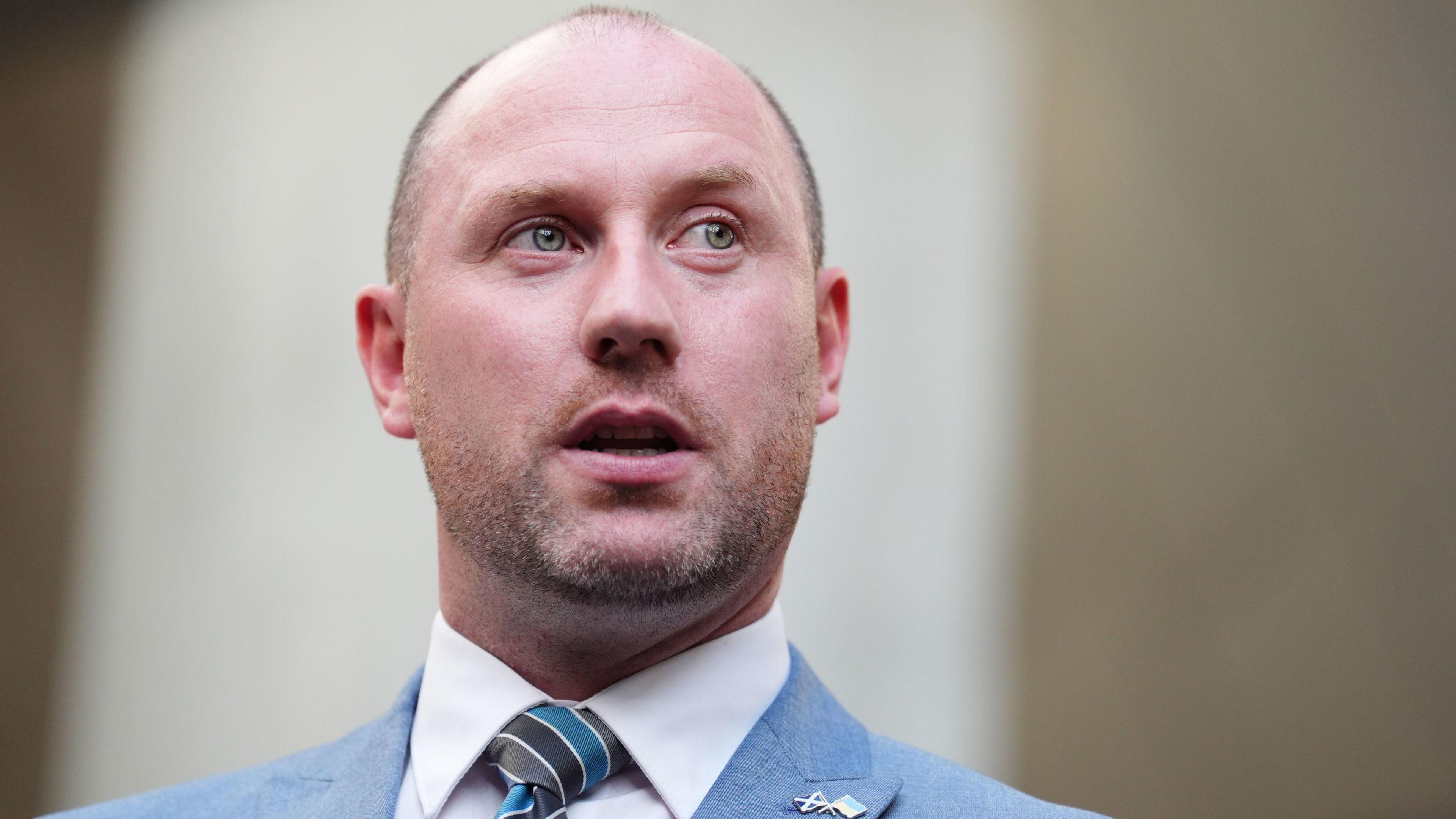 A man standing, wearing a pale blue suit. He is balding, with stubble.