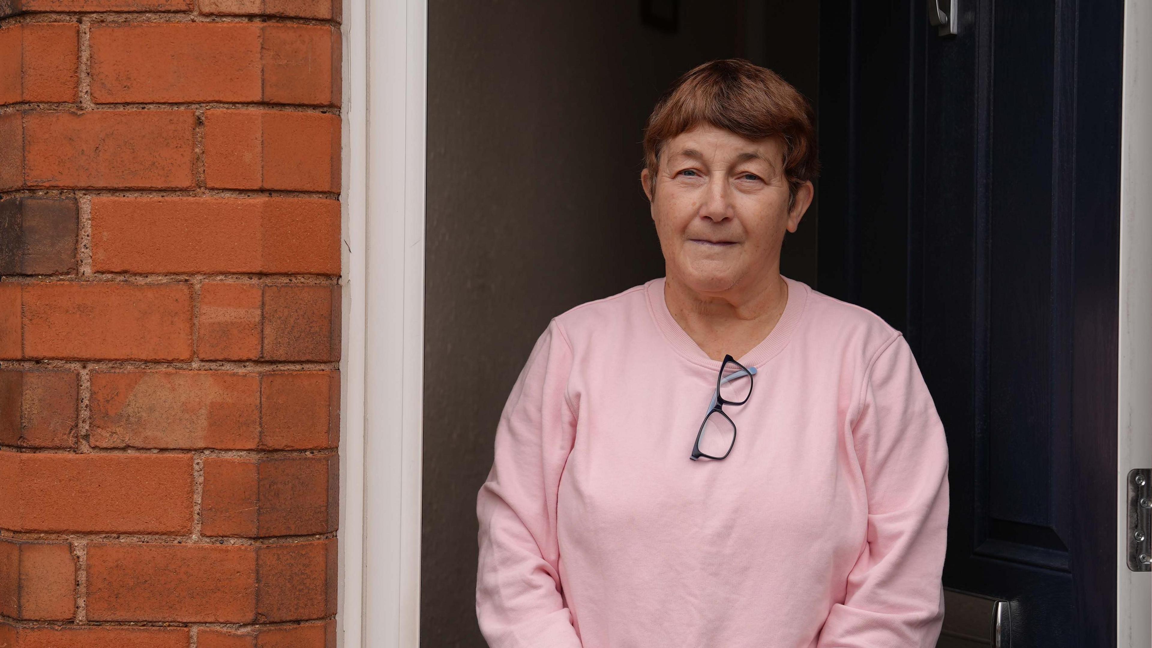 A woman in a light pink jumper with her glasses tucked into the top. She has short, brown hair and is standing in front of her dark-coloured front door, which is partially open