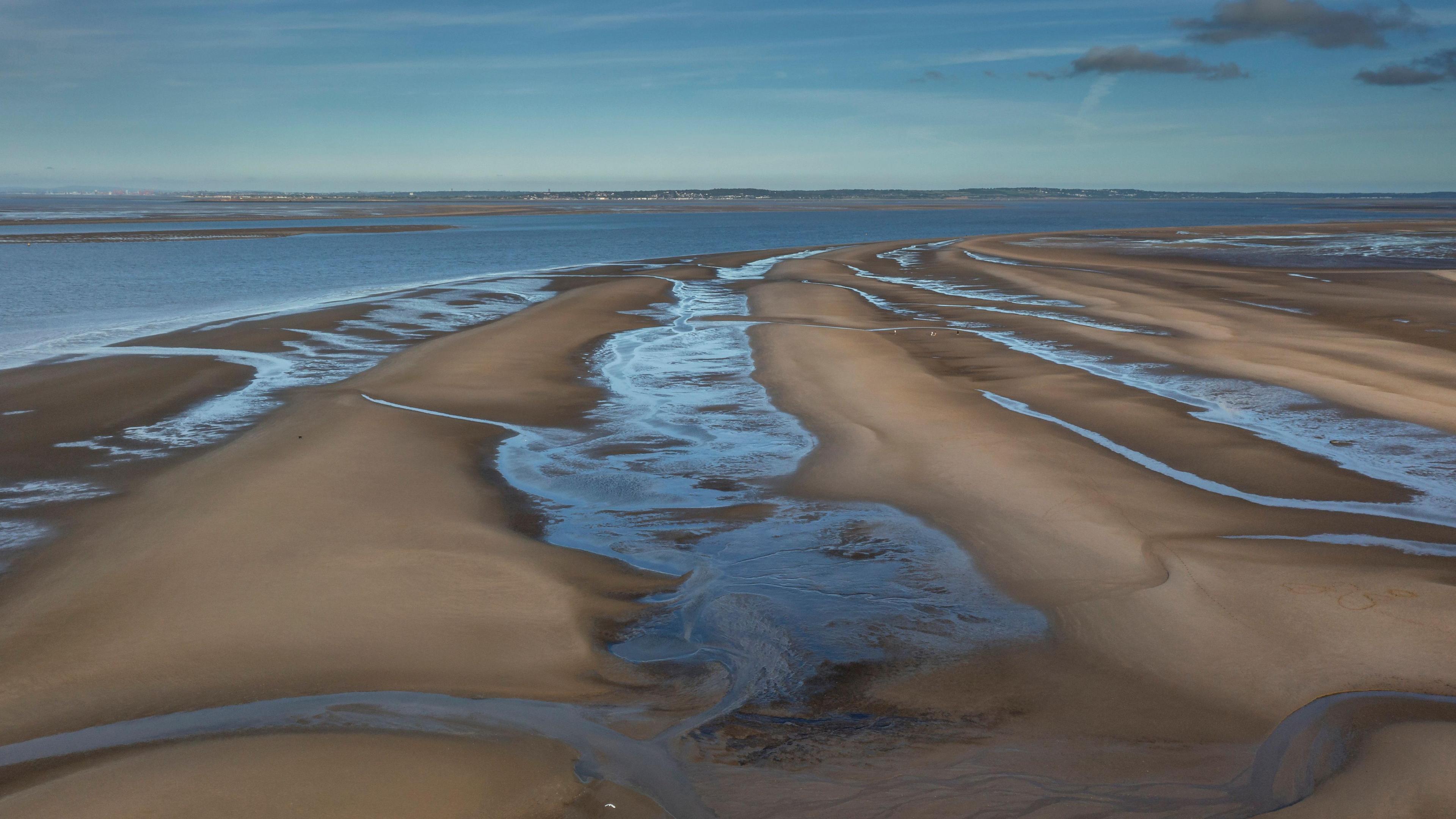 An aerial view of Talacre Beach and the Dee Estuary. There are bands of sand and water in channels running parallel to each other with the estuary beyond. In the horizon the other side of the estuary can just be seen, with a wide blue sky above.