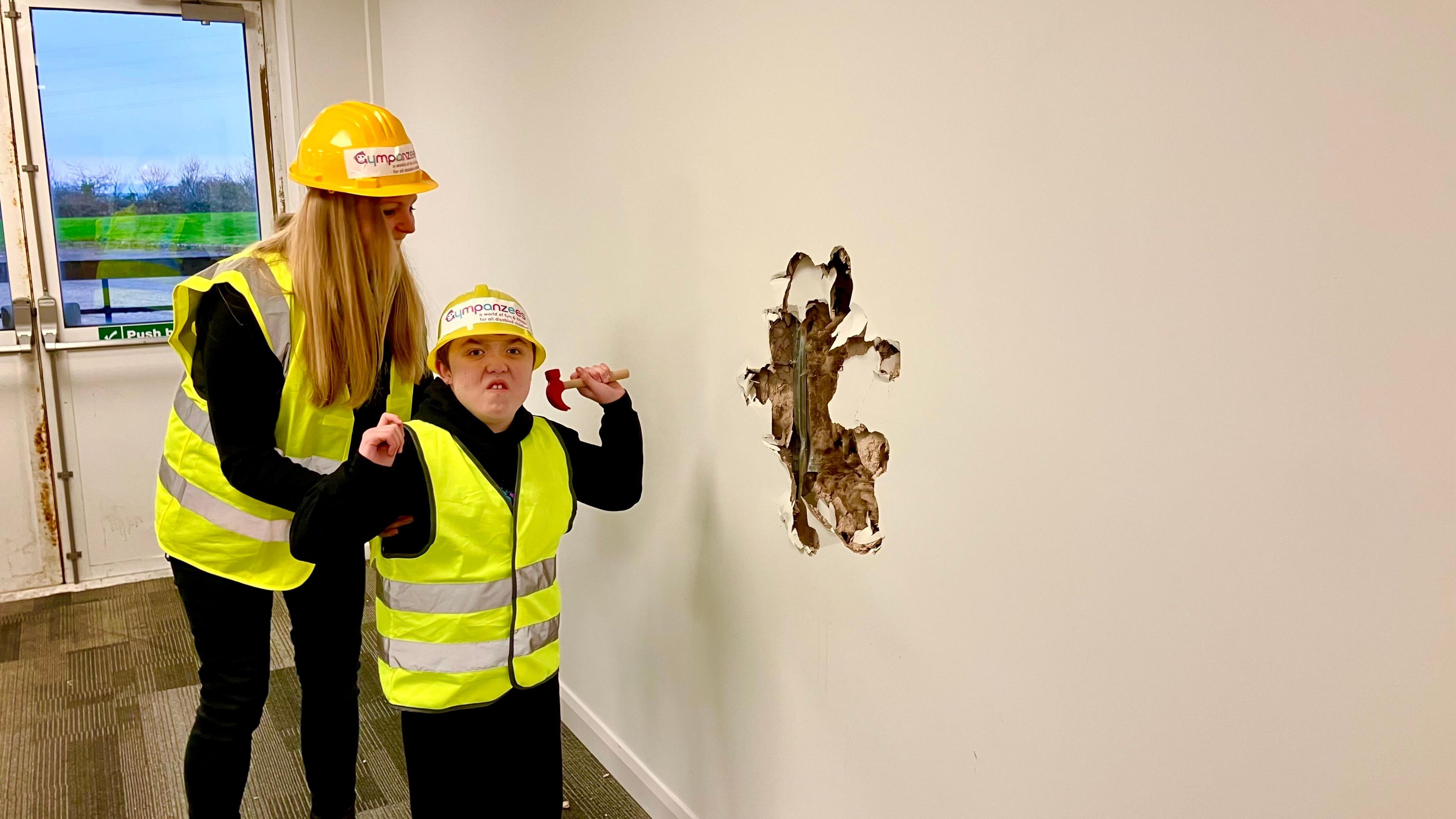 Karina Kay with her son Joel standing next to a wall with a hole in it, pretending to have carried out the work, with Joel hold a small toy hammer.