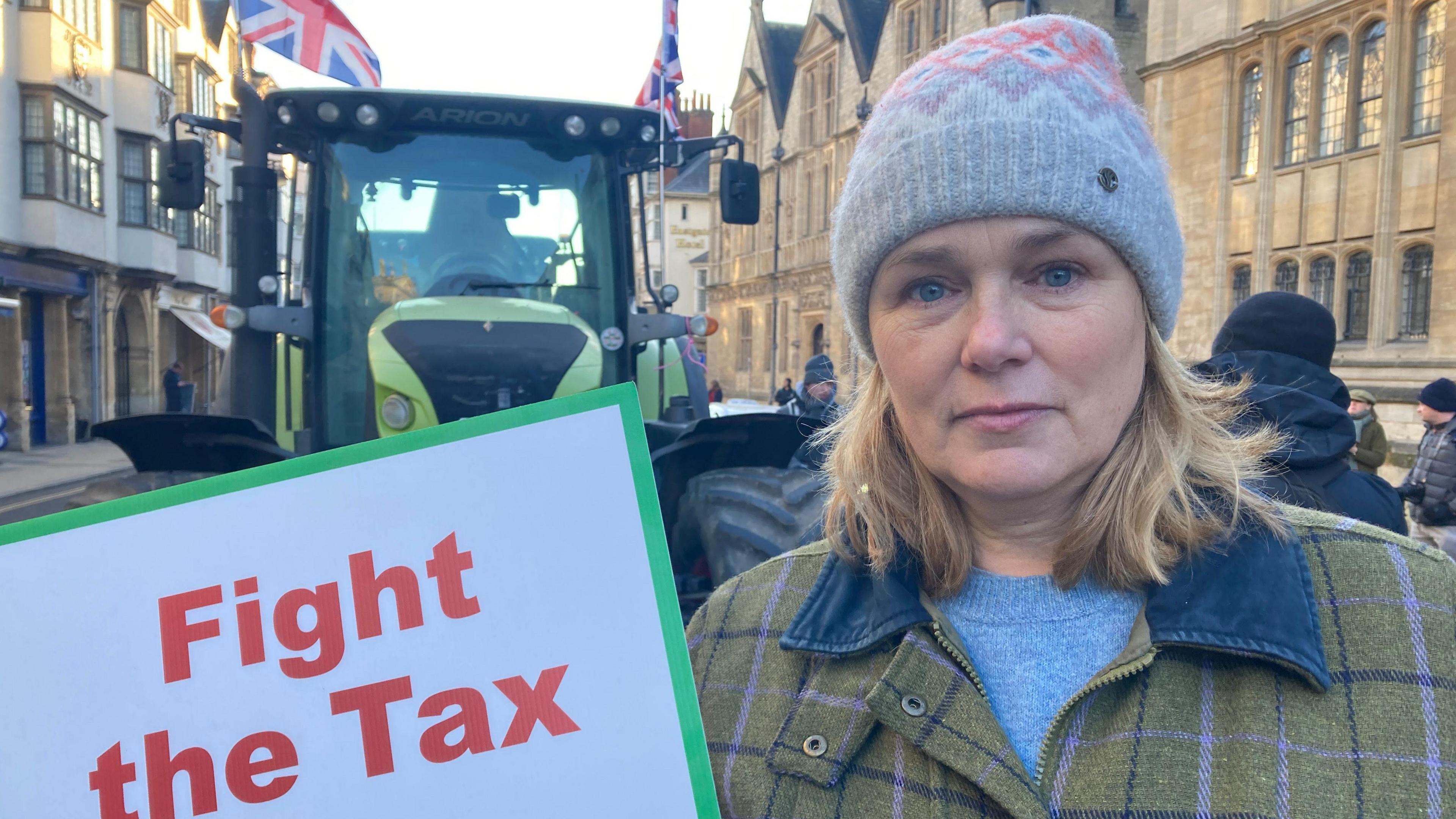 Heidi Smith wearing a tweed coat and woolly hat is holding a sign saying 'Fight The Tax'. A tractor flying Union Jack flags is behind her.