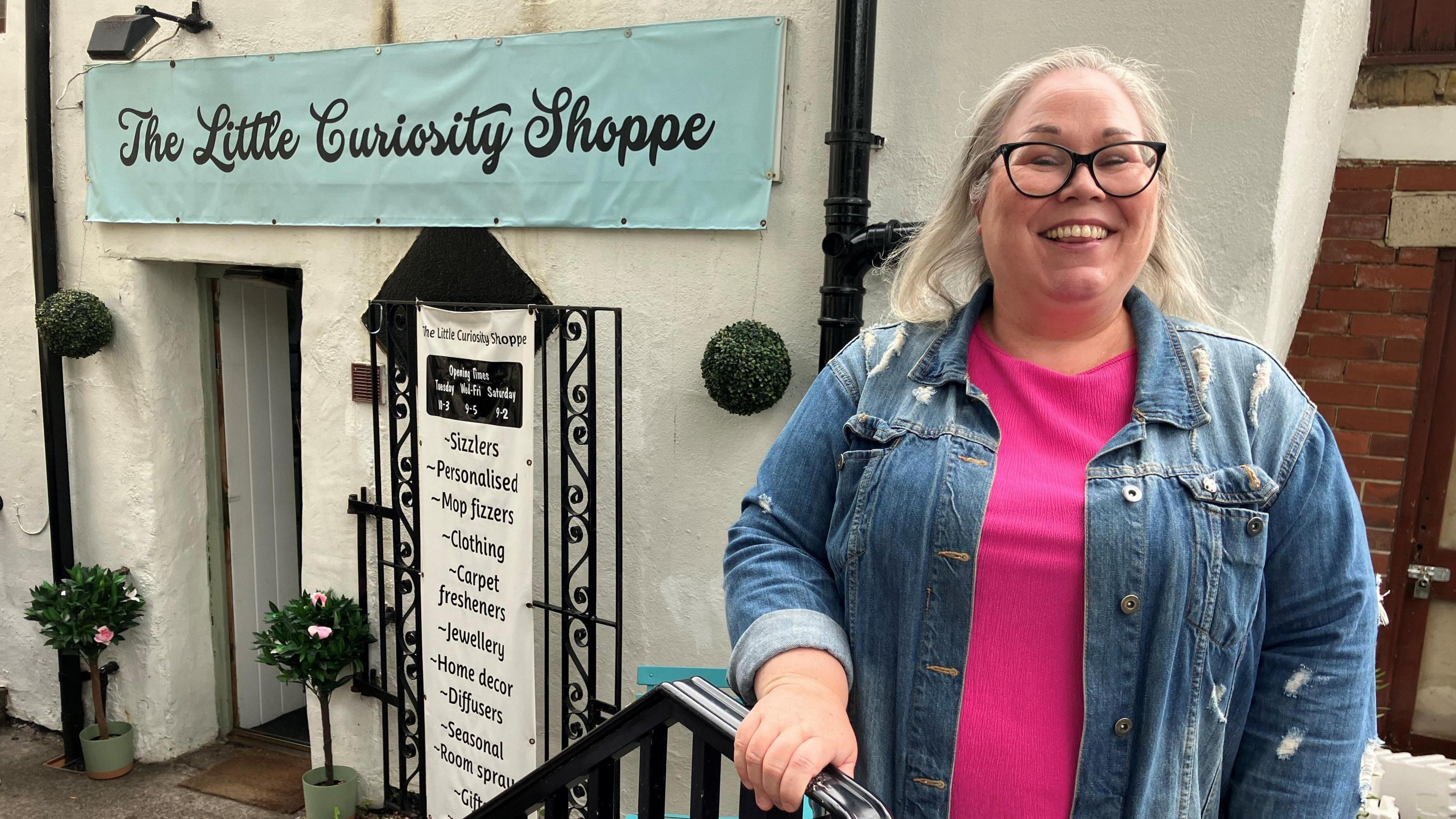 A blond, bespectacled woman stands in front of a shop. She is wearing a pink top and denim jacket over it as she holds on to a railing. A banner above the shop entry reads "The Little Curiosity Shoppe".