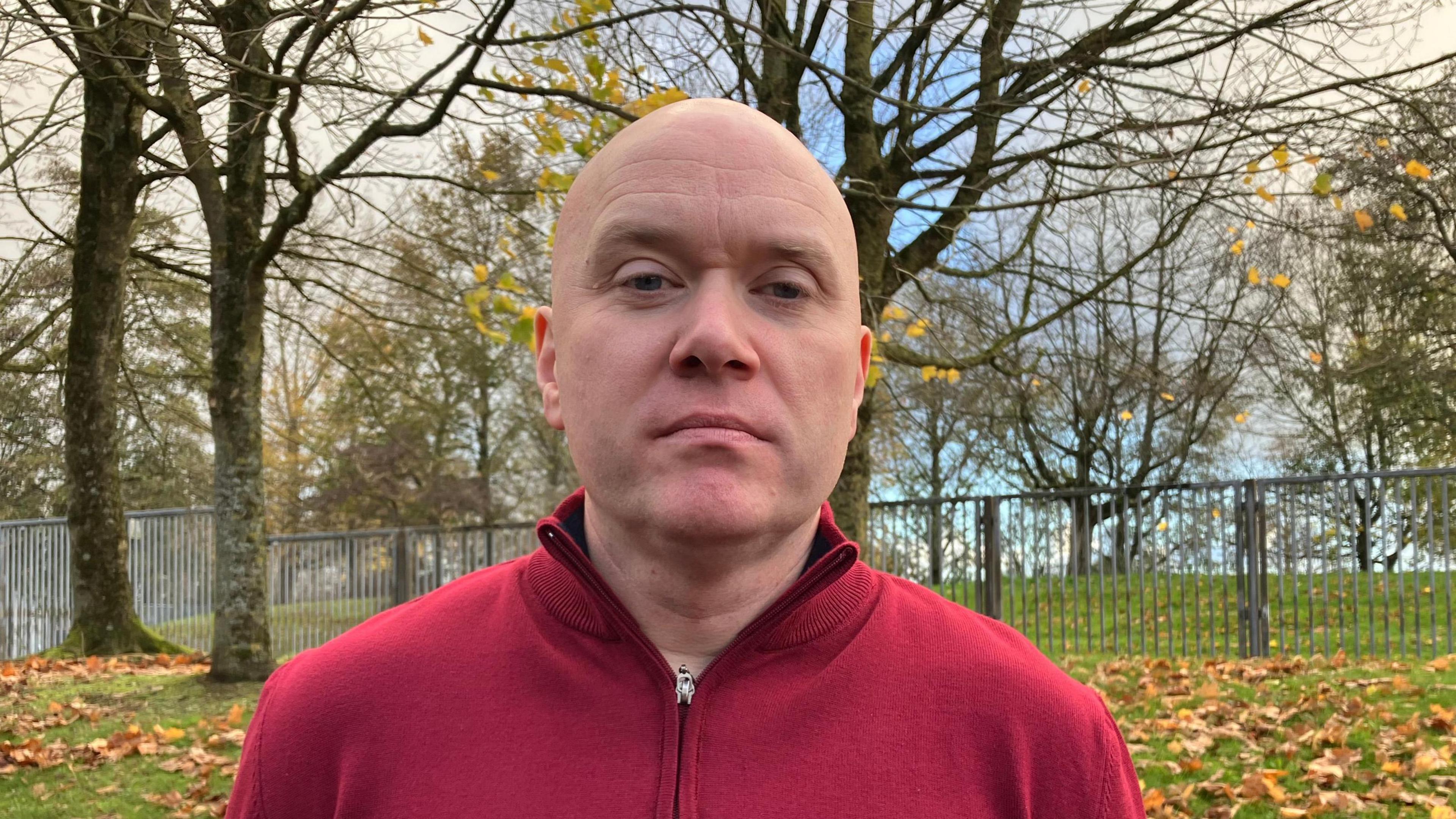 A bald headed man wearing a zip up red jumper. Behind him is trees, grass and a metal fence with brown leaves on the ground.