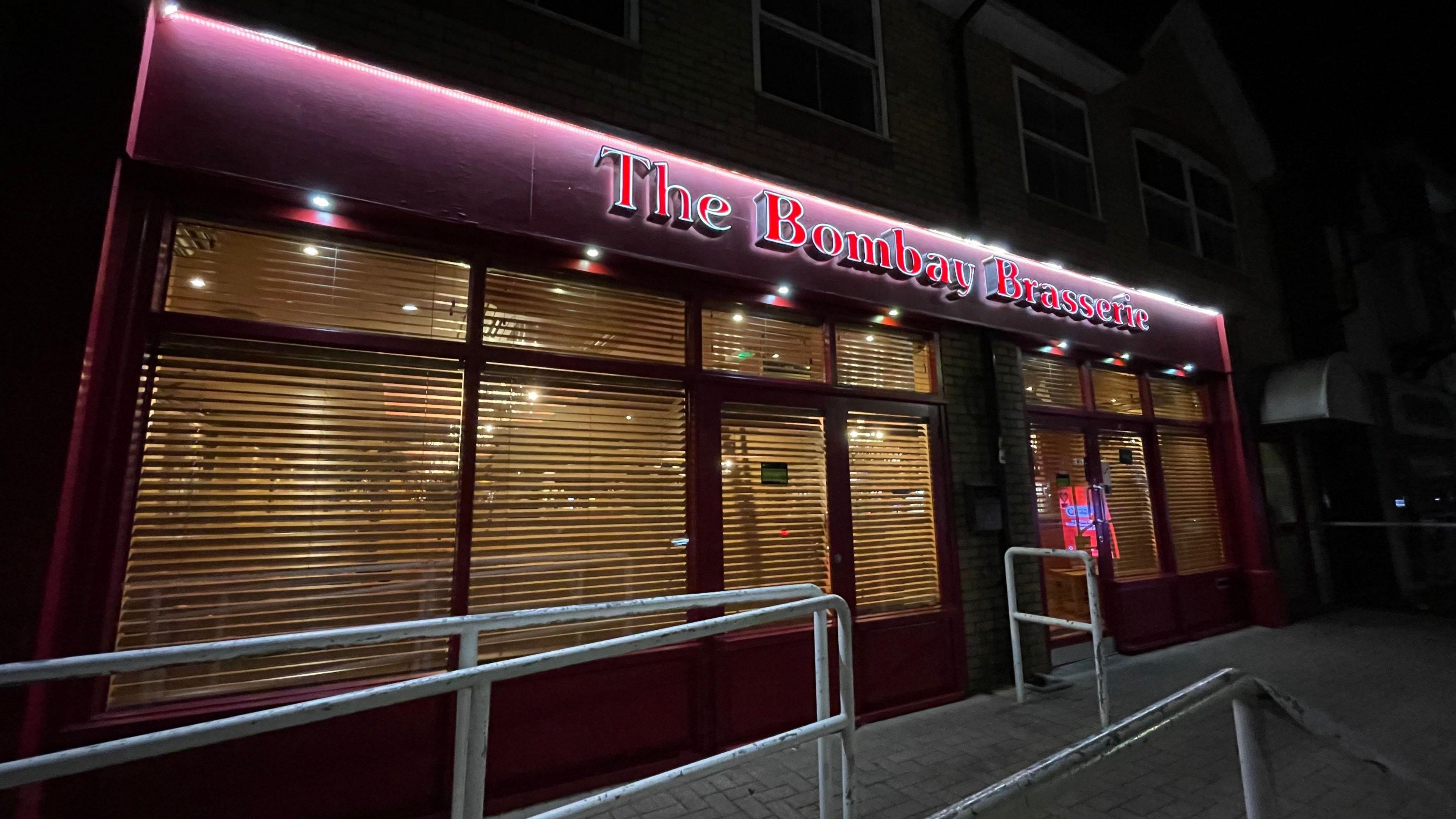 An exterior shot of the Bombay Brasserie. The front of the restaurant is painted purple, with the letters of The Bombay Brasserie written in red and back-lit. There are several windows, which all have blinds over them, through which yellow light is seeping out.