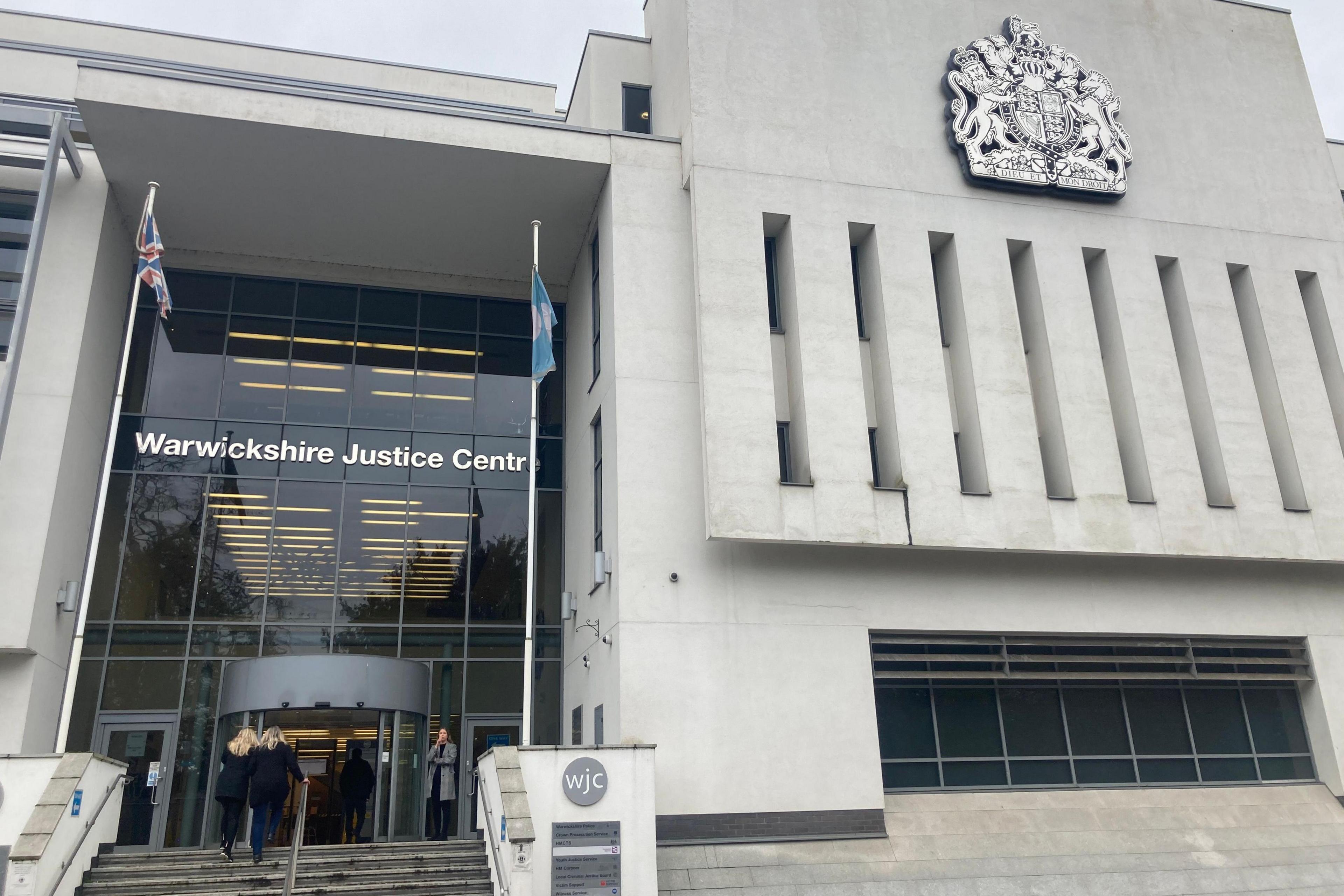 An image of Warwickshire Crown Court, a grey stone building with glass frontage flanked with flags, with people at the top of some steps