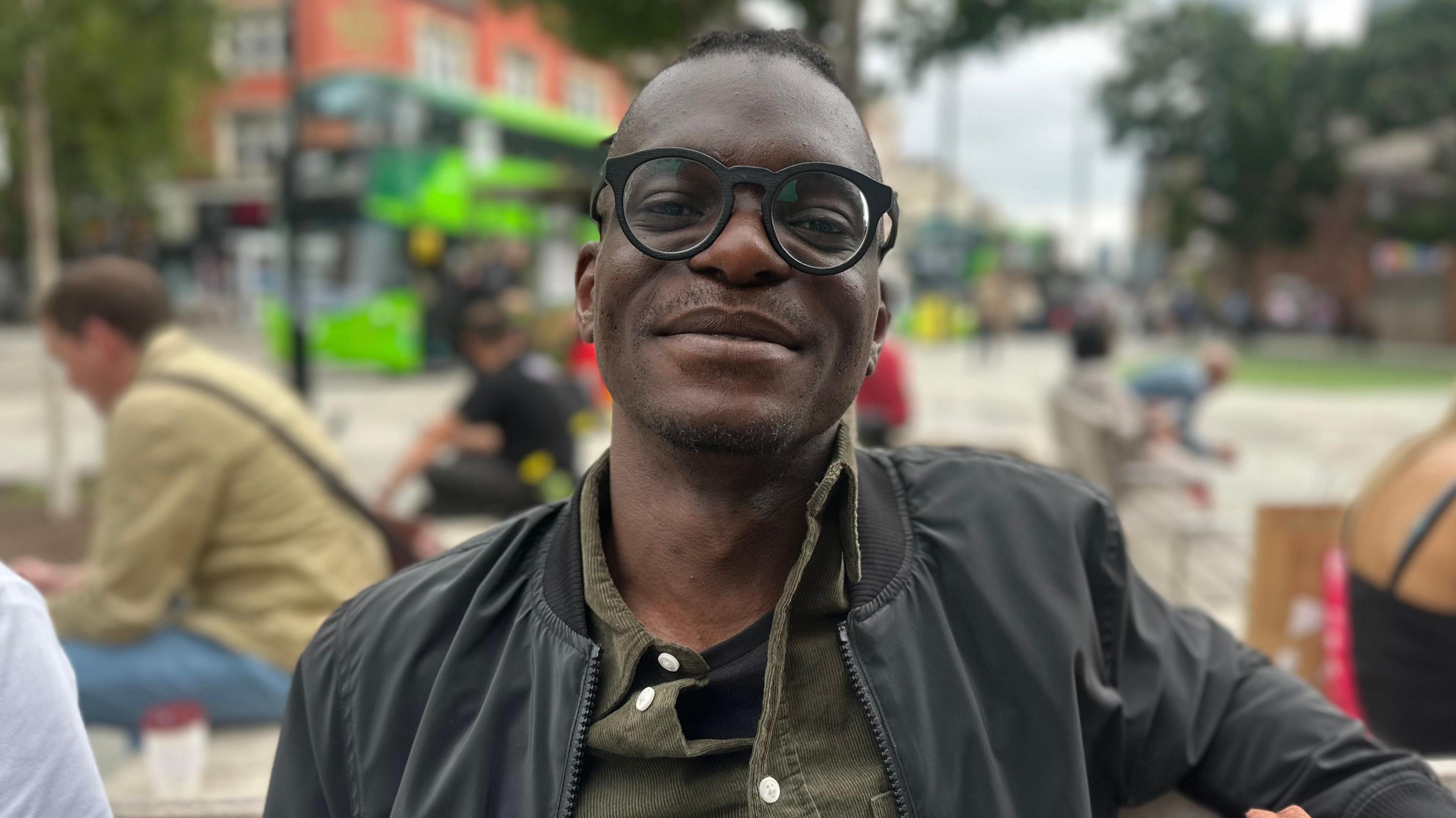 A young man with large round spectacles and a dark coat. He is sat on a city centre bench.