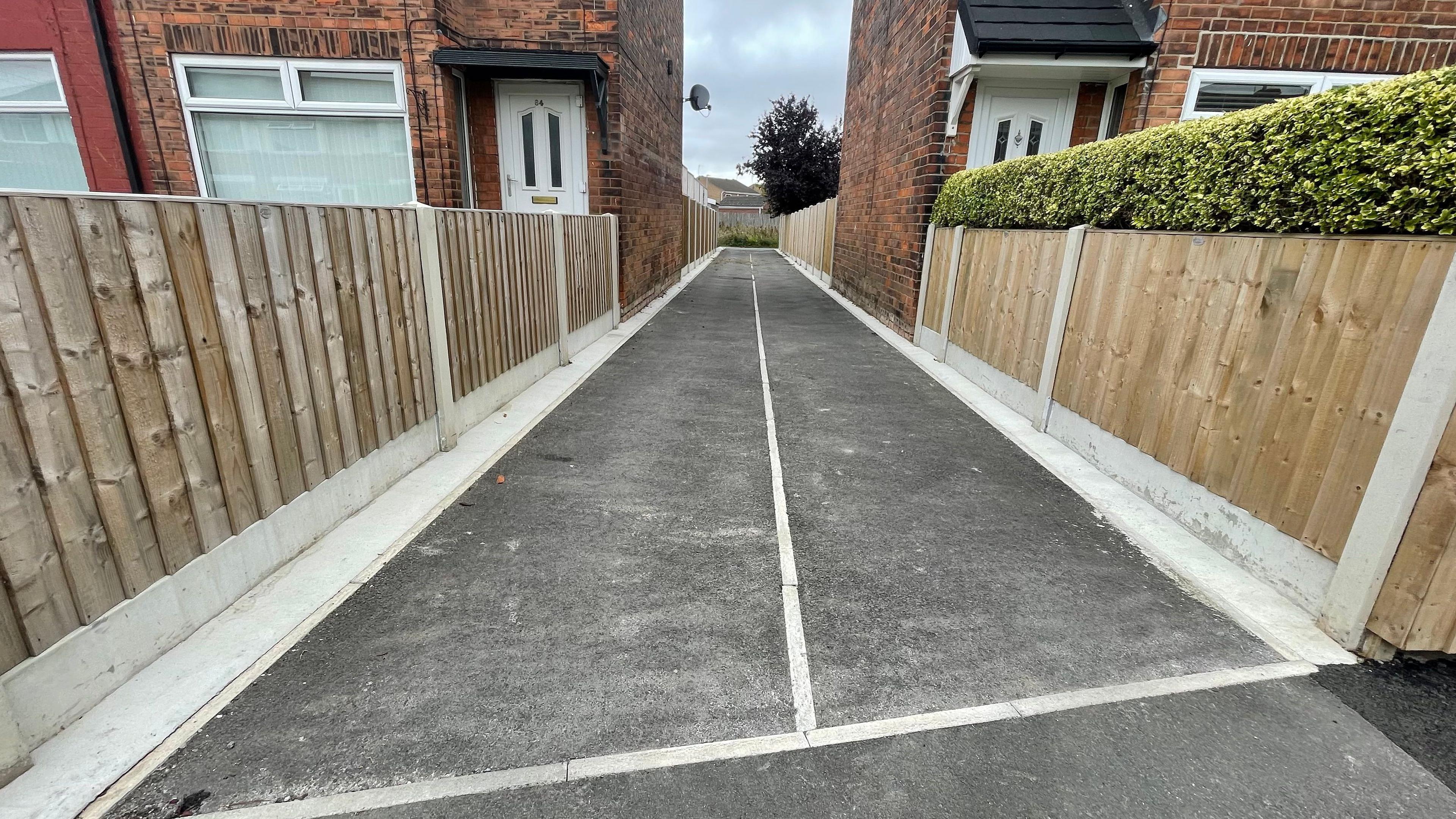 A newly resurfaced tarmac alleyway stretches into the distance between wooden fences and two red brick houses.